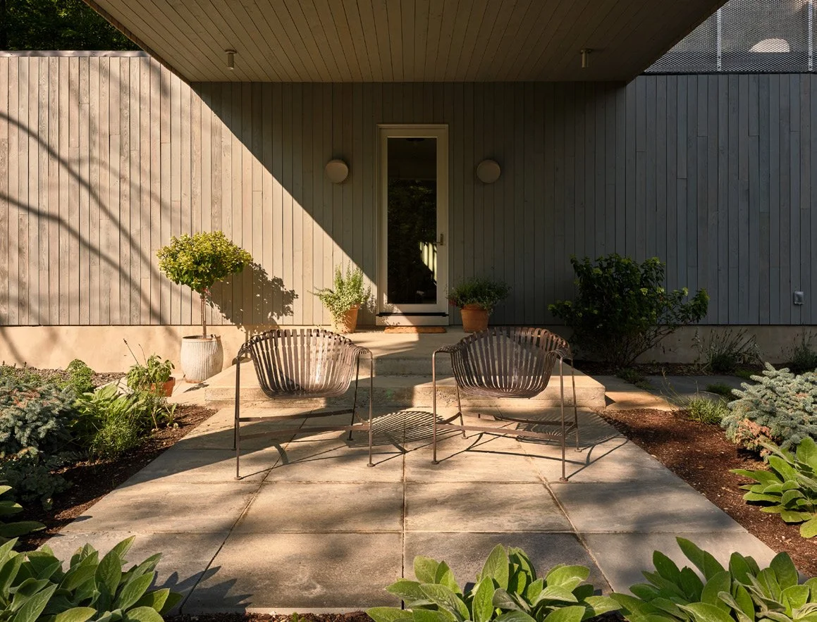 A modern backyard patio with two metal chairs and a small table, surrounded by lush greenery and potted plants, in front of a house with gray wooden siding and a glass door.