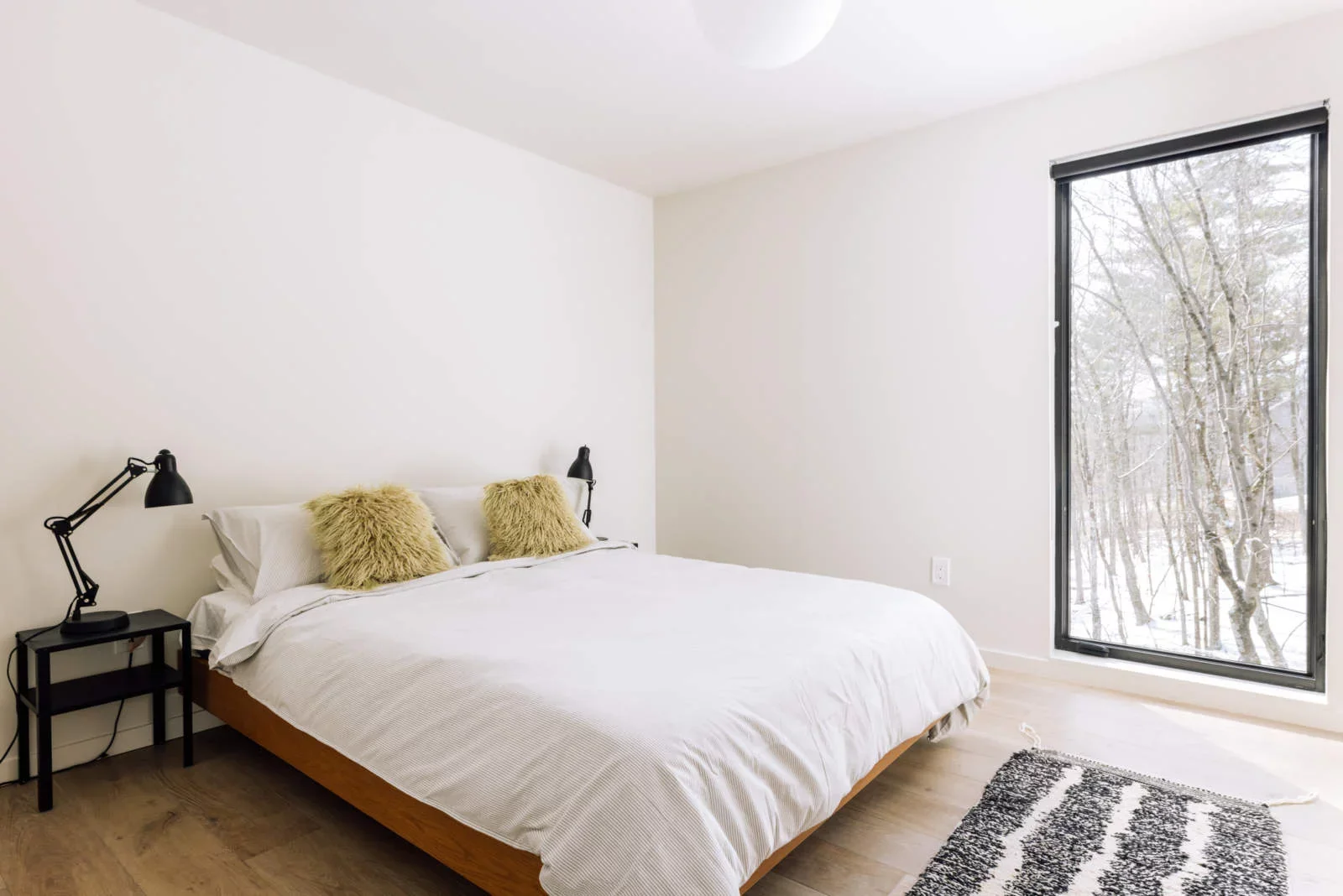 A bedroom with a white bedspread, two beige fluffy pillows, black bedside tables, and black adjustable lamps. There's a large window showing a snow-covered tree outside, and a black and white rug on light wood floors.