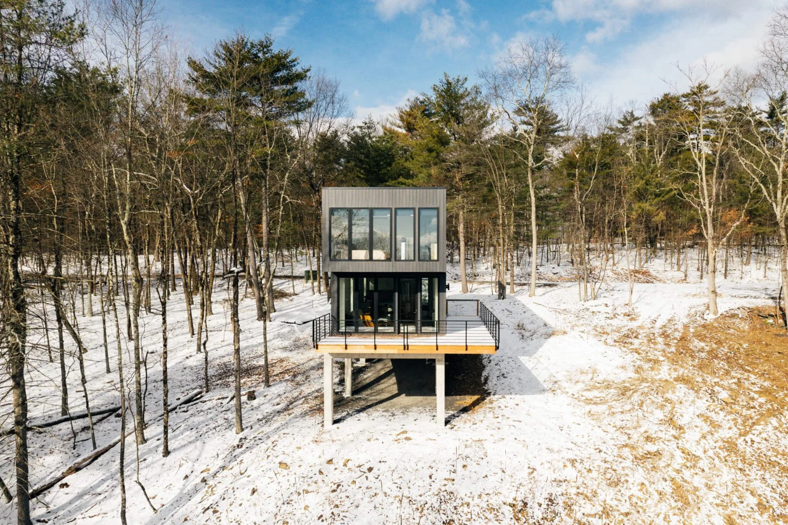 Modern two-story house elevated on stilts in a snowy wooded landscape with clear sky.