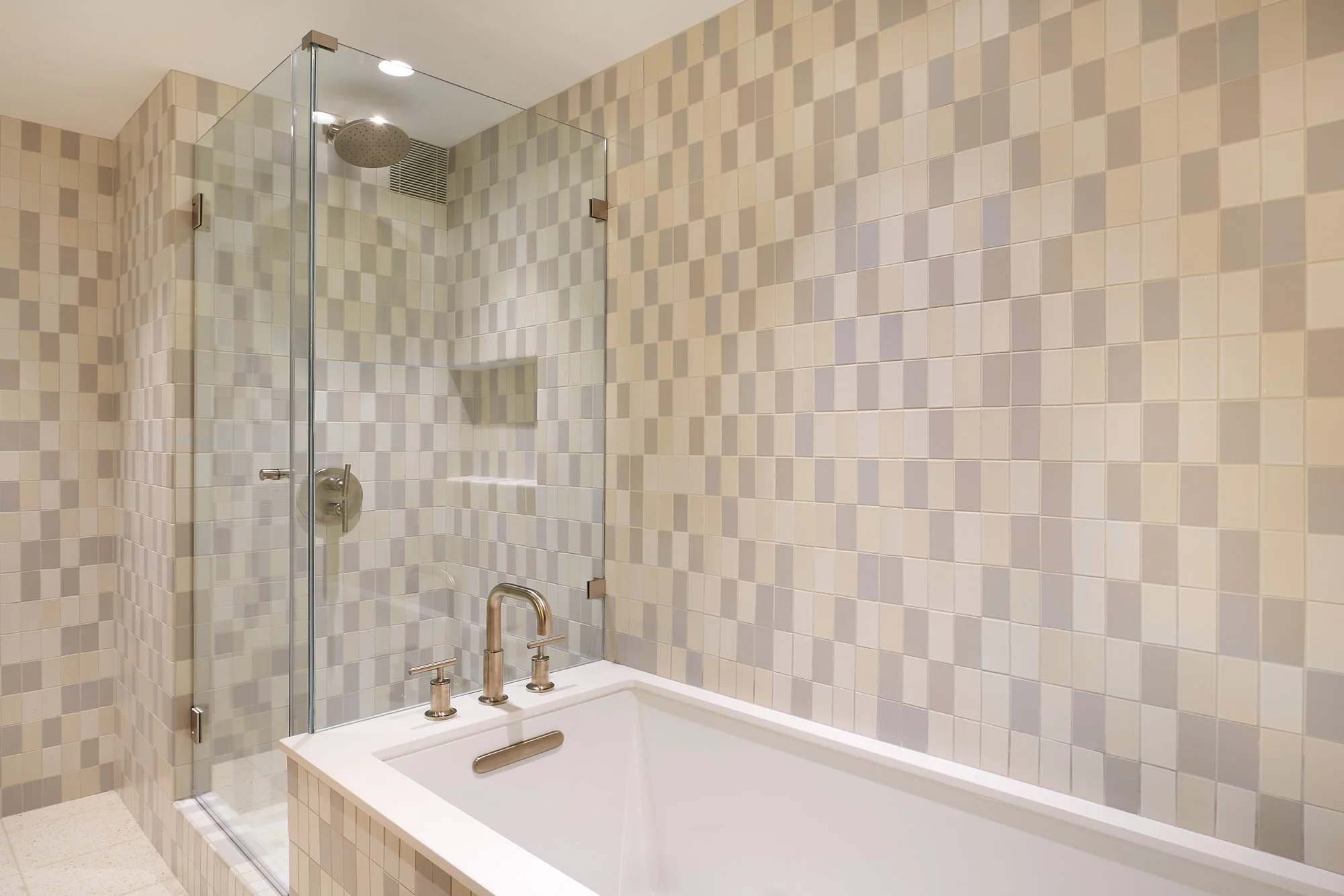 Bathroom with a glass-enclosed shower area with a rainfall showerhead and a built-in niche, and a white bathtub with silver fixtures, all surrounded by beige and cream mosaic tiles.