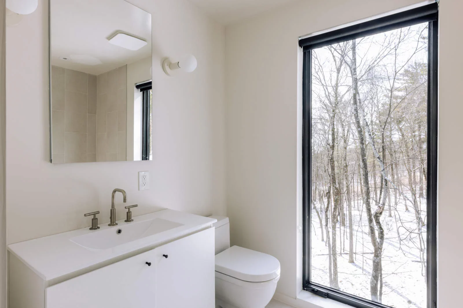Bathroom with a white sink, mirror, and toilet, next to a large window showing a snowy outdoor scene with trees.