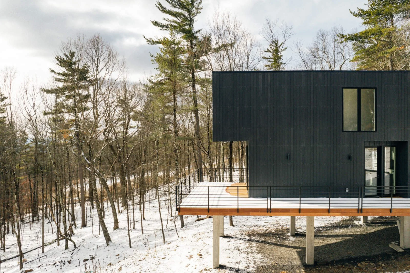 A modern black house on stilts with a wooden deck, set in a snowy wooded area, with leafless trees and cloudy sky in the background.