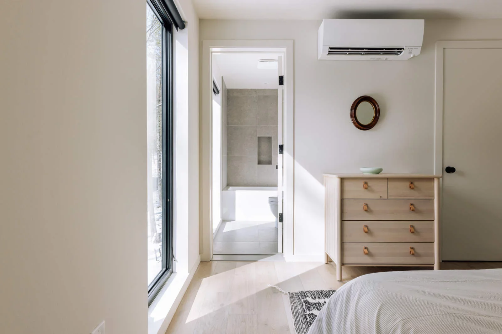 A bedroom with a sliding glass door, a wooden dresser, small mirror, and an air conditioner. An open door reveals a bathroom with a tiled shower and a toilet.