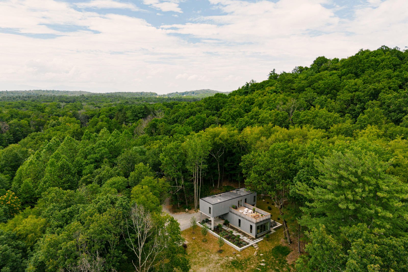 A modern house surrounded by dense green trees and forest, with a gravel driveway leading to the house, under a partly cloudy sky.