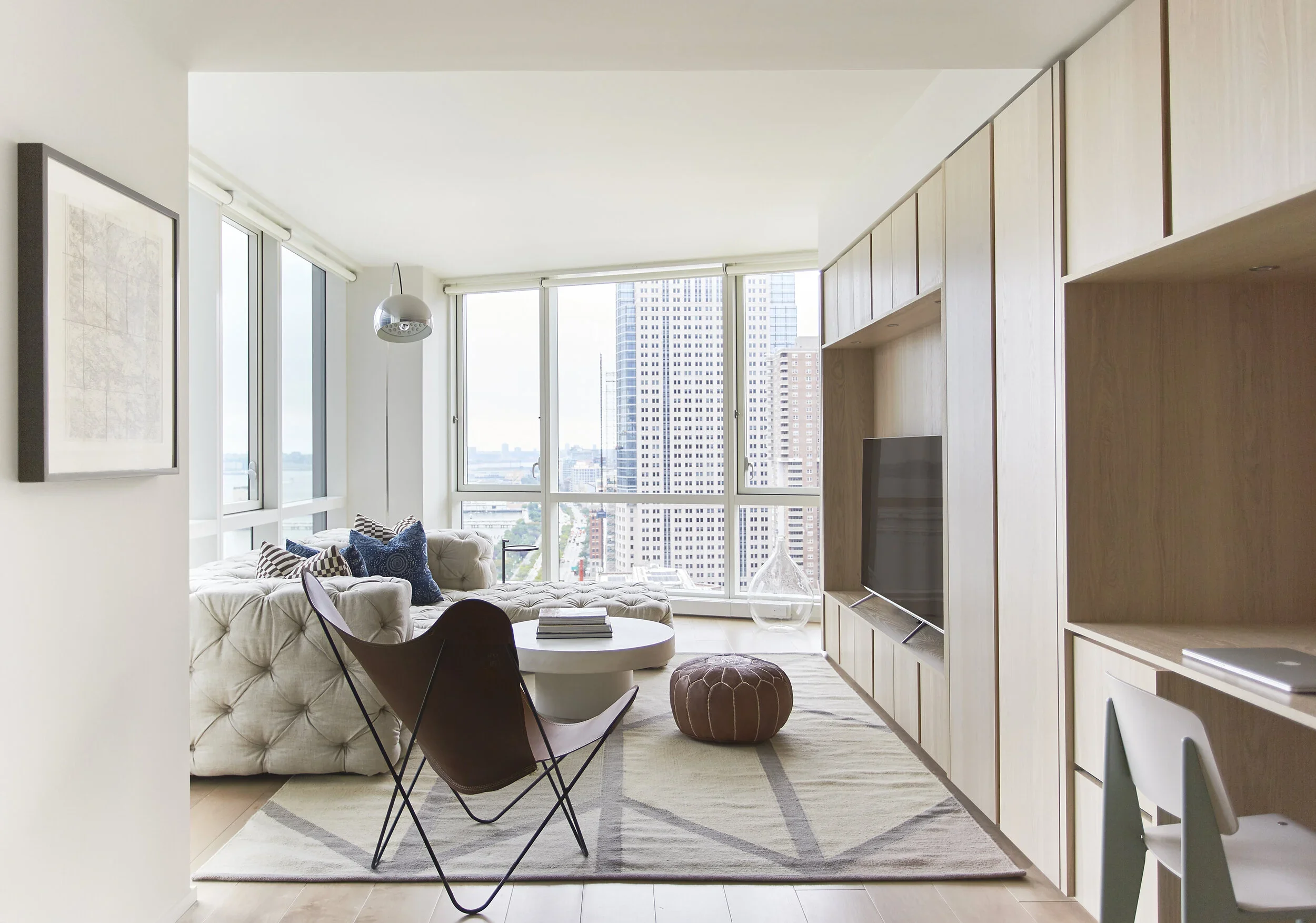 Modern living room in high-rise apartment with large windows, beige tufted sofa with blue pillows, round coffee table, patterned rug, brown leather butterfly chair, wooden wall unit with TV, and cityscape view.