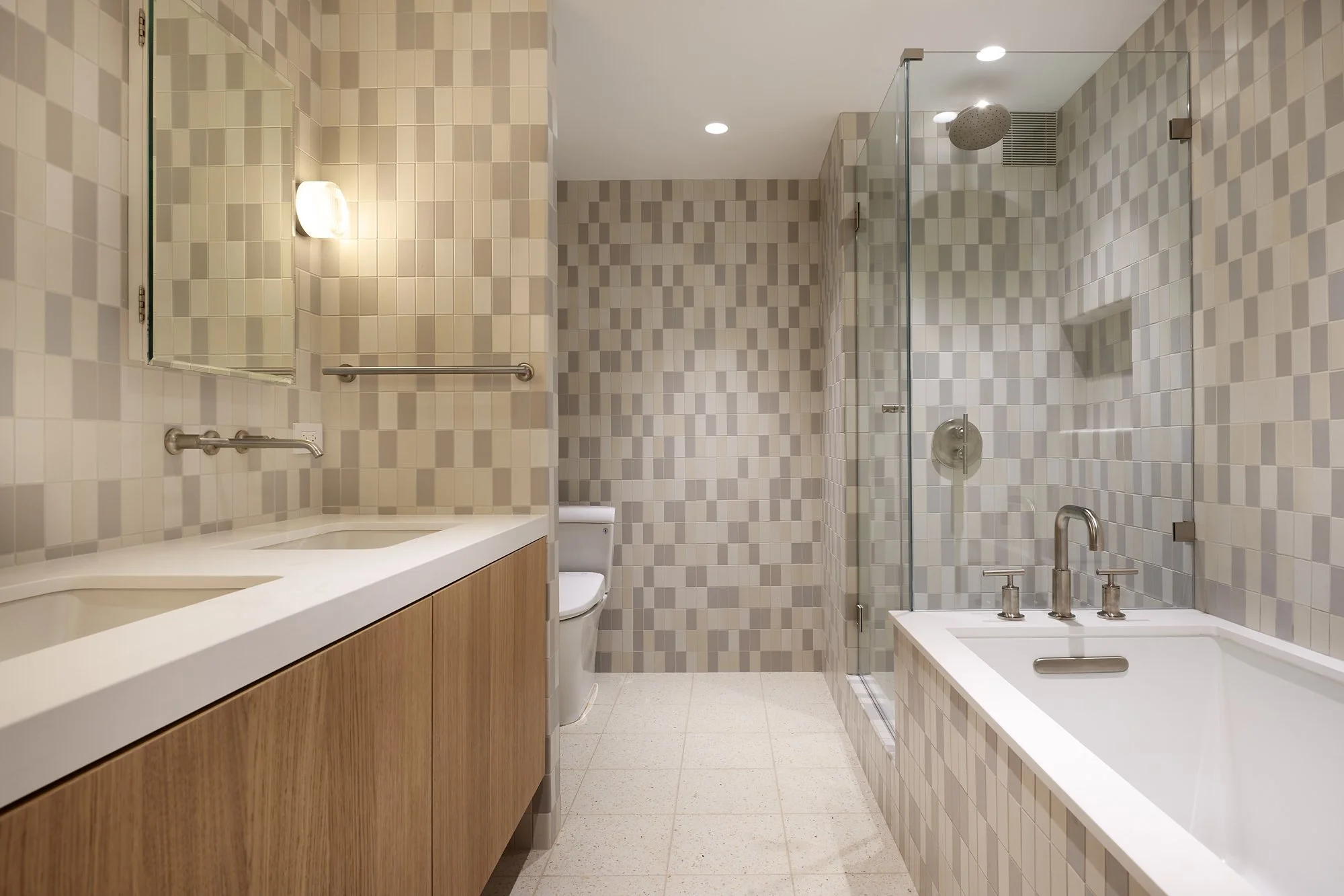 Modern bathroom with beige tile walls, white countertop with dual sinks, wooden cabinet, wall mirror, toilet, glass shower with rain showerhead, bathtub, and metallic fixtures.