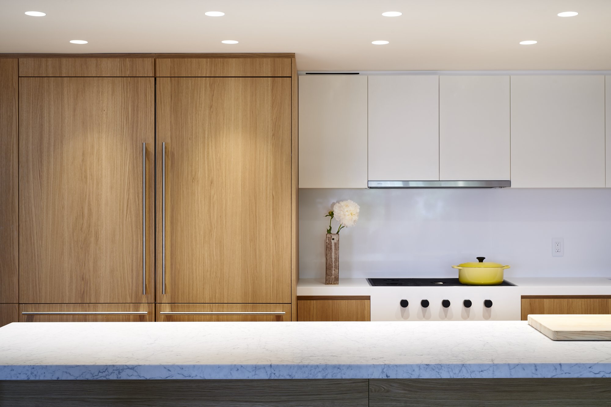 Modern kitchen with wooden cabinets, white upper cabinets, yellow pot on stove, vase with white flowers, and marble countertop.