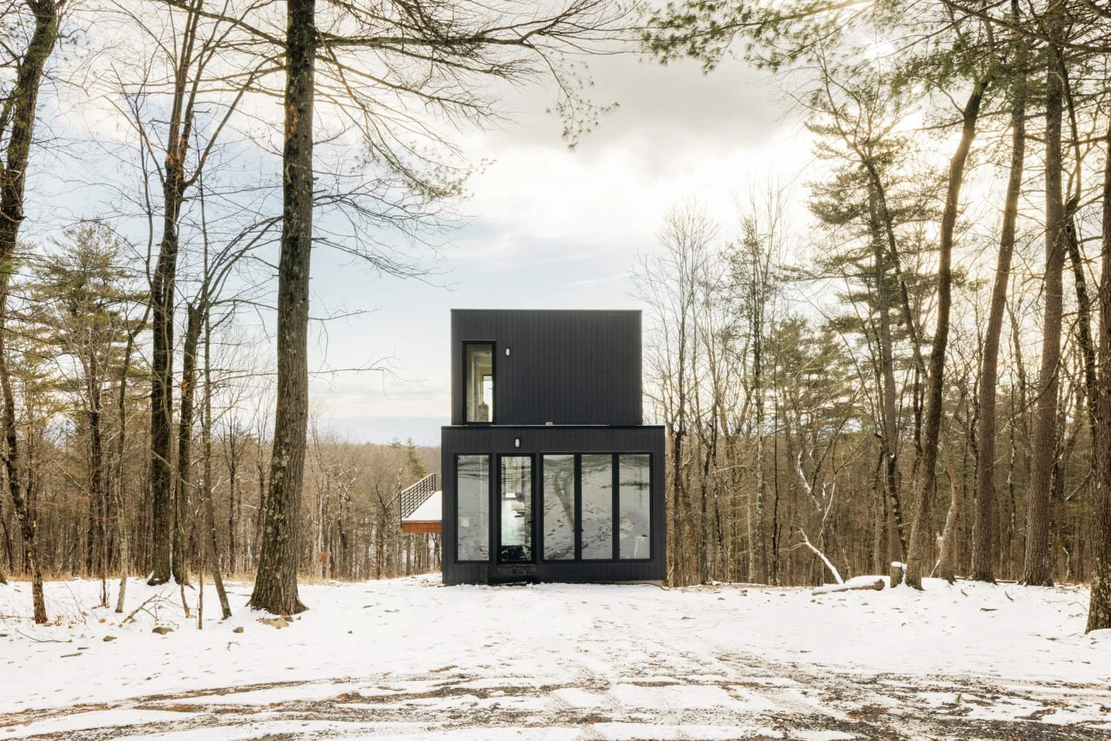 A modern two-story black house with large glass windows on the ground floor, set in a snowy forest clearing surrounded by tall, leafless trees under a cloudy sky.