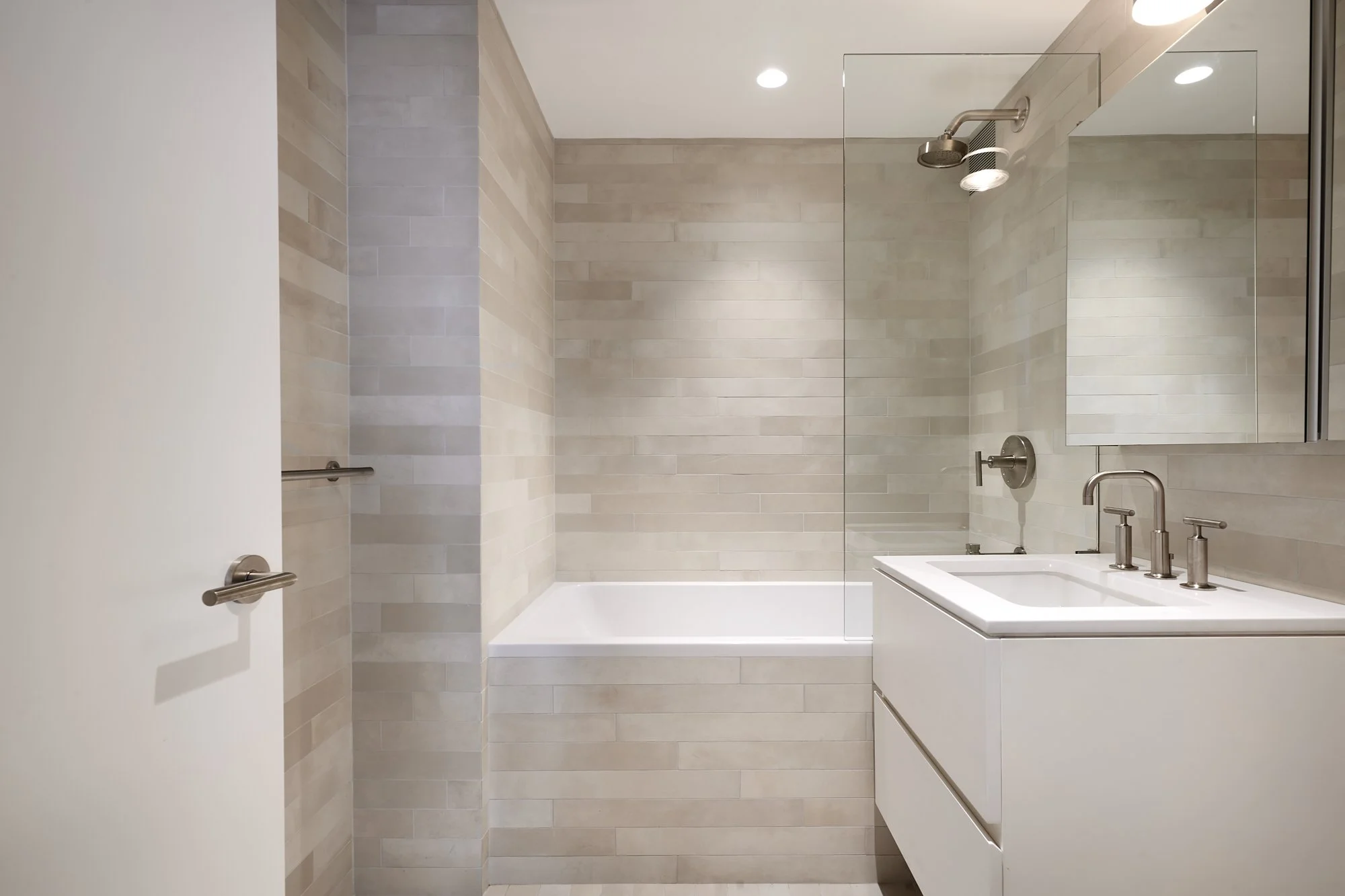 Modern bathroom with beige tiled walls, white bathtub, glass shower divider, and white vanity with stainless steel fixtures.