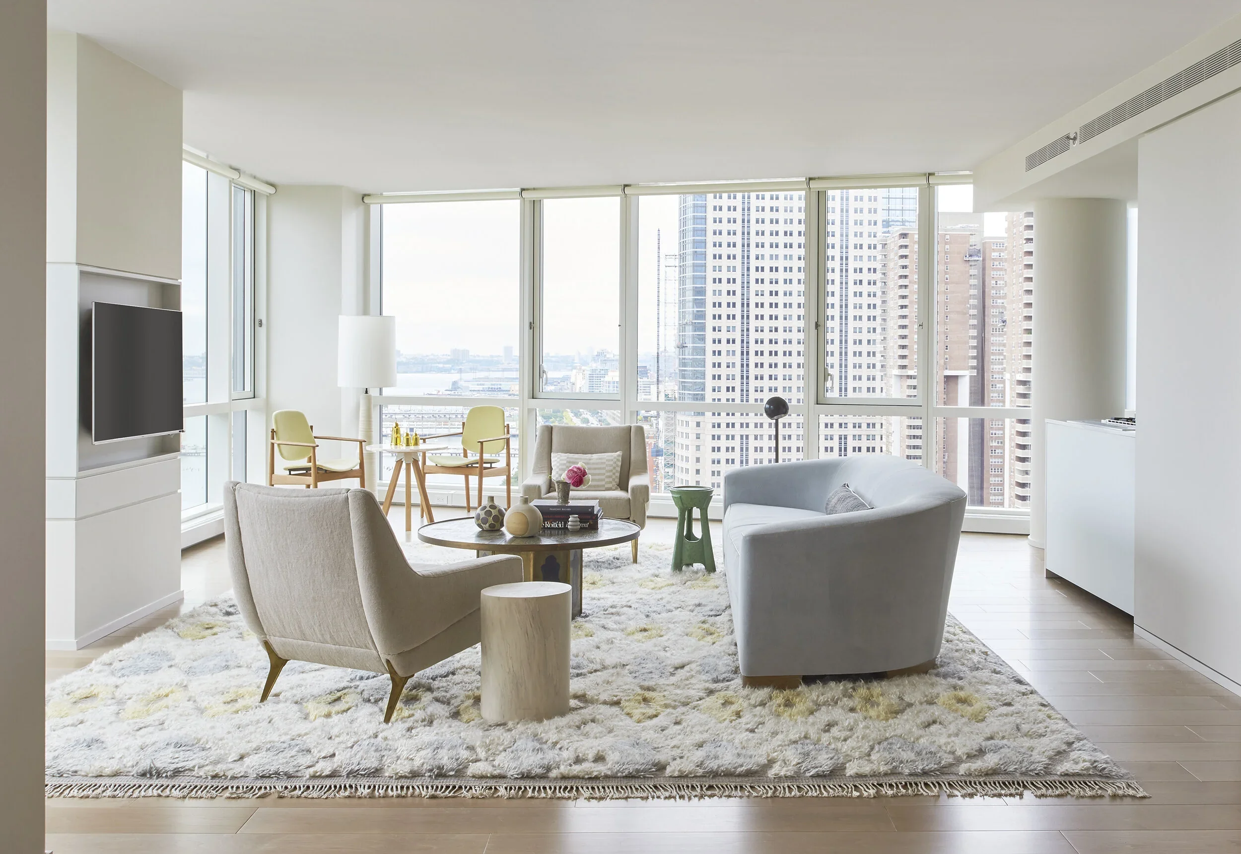 Modern living room with large windows, white walls, beige and light gray furniture, a shag rug, and a cityscape view.