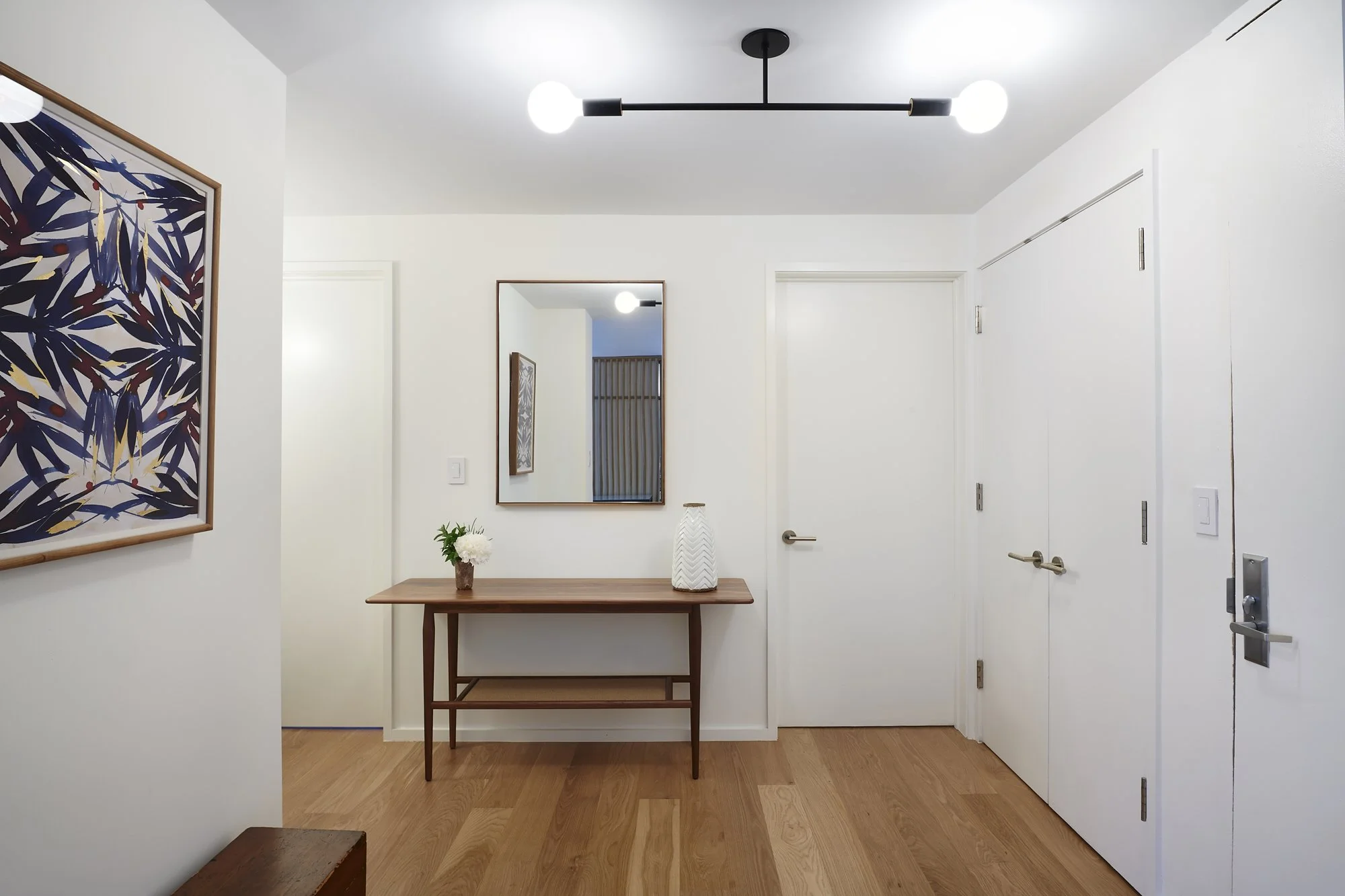 Modern entryway with white walls, wooden flooring, a wooden console table, a mirror, decorative vase, and framed artwork, with a contemporary ceiling light fixture.
