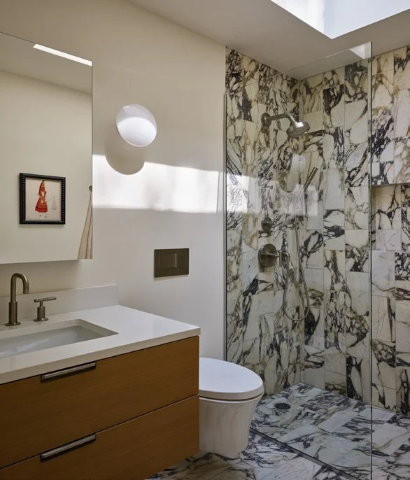 Bathroom with a white sink, wooden vanity, mirror, wall light, toilet, and a marble shower area with a showerhead and glass door.