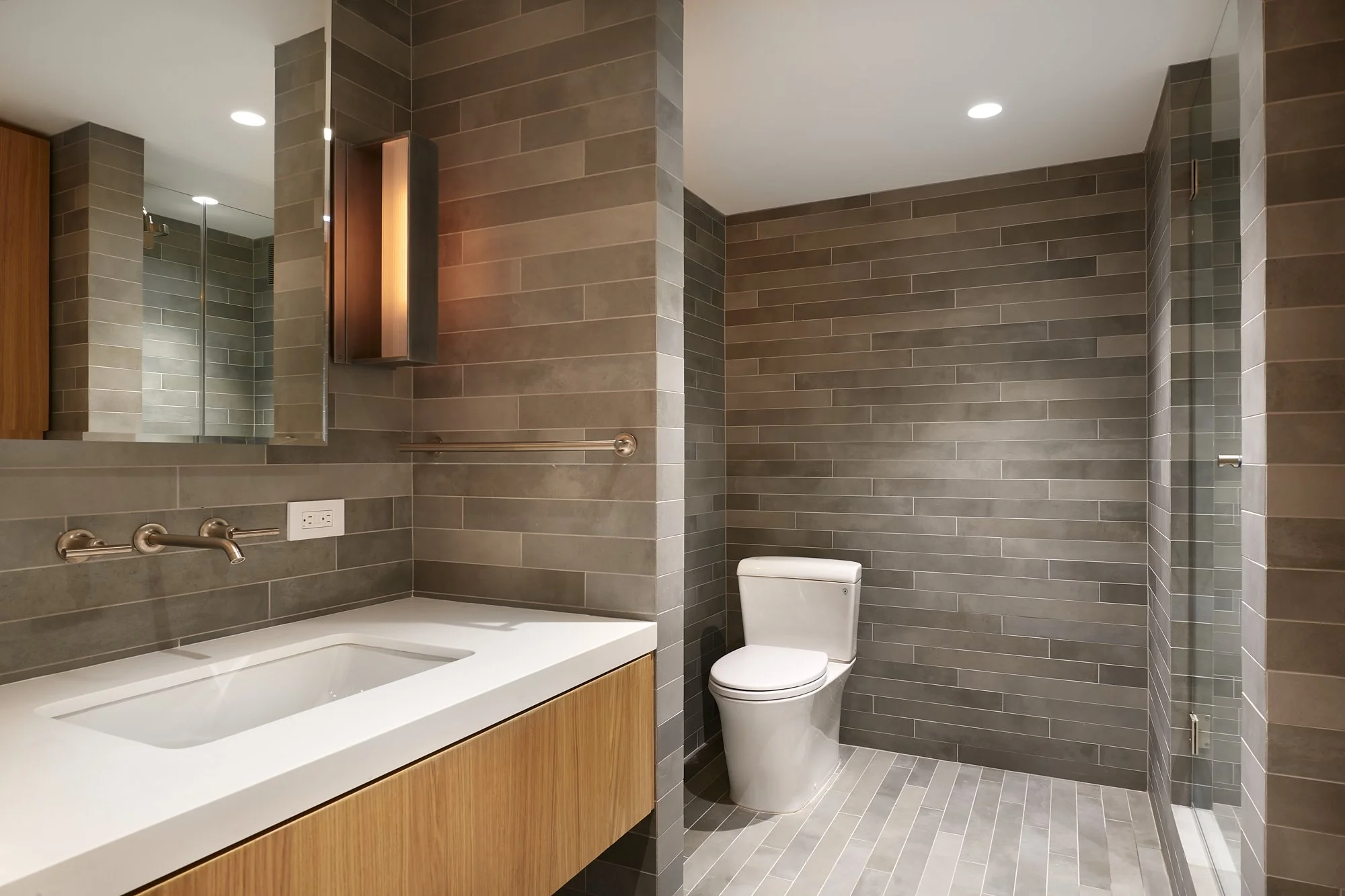 Modern bathroom with grey tiled walls and floor, white toilet, wooden vanity with white countertop, single sink, large mirror, and a glass-enclosed shower area.
