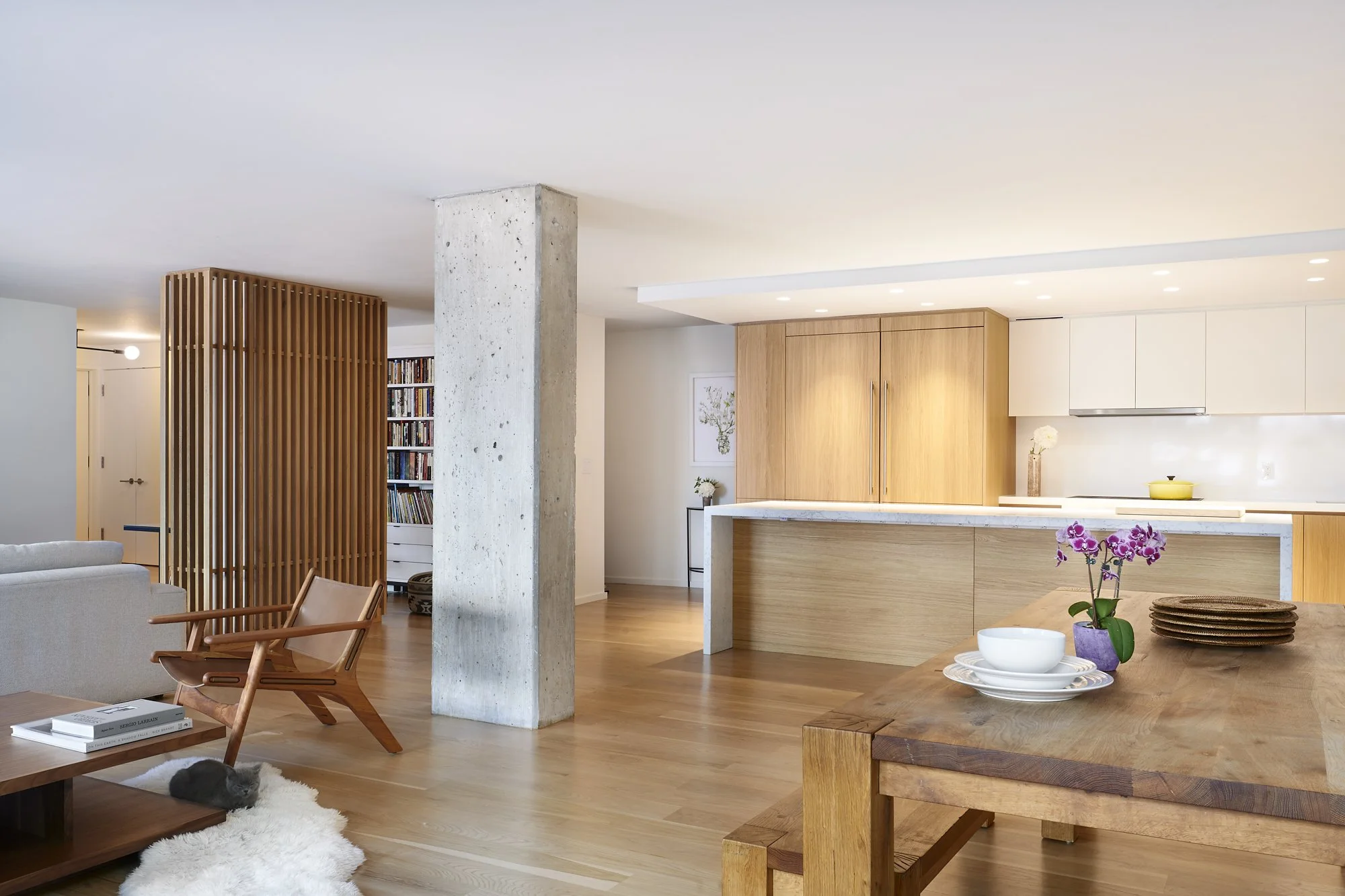 Open-concept living room and kitchen with wooden flooring, white and wood cabinets, and a concrete column. A wooden dining table with dishes and a pink orchid, a lounge chair, and a shaggy rug with a cat are visible.