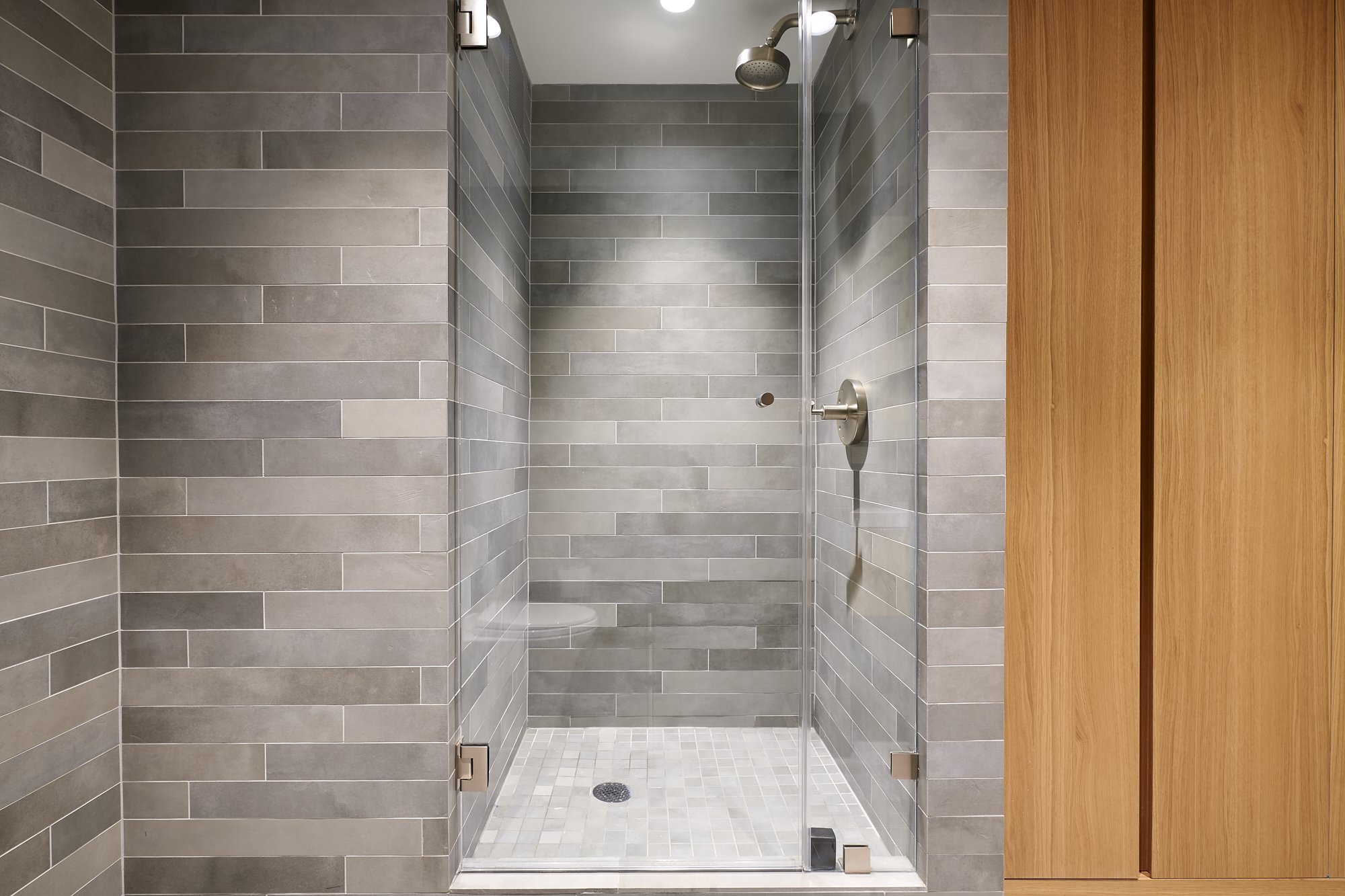 Modern glass shower with gray tile walls and small square white and gray tiled floor, partially open wooden door on the right