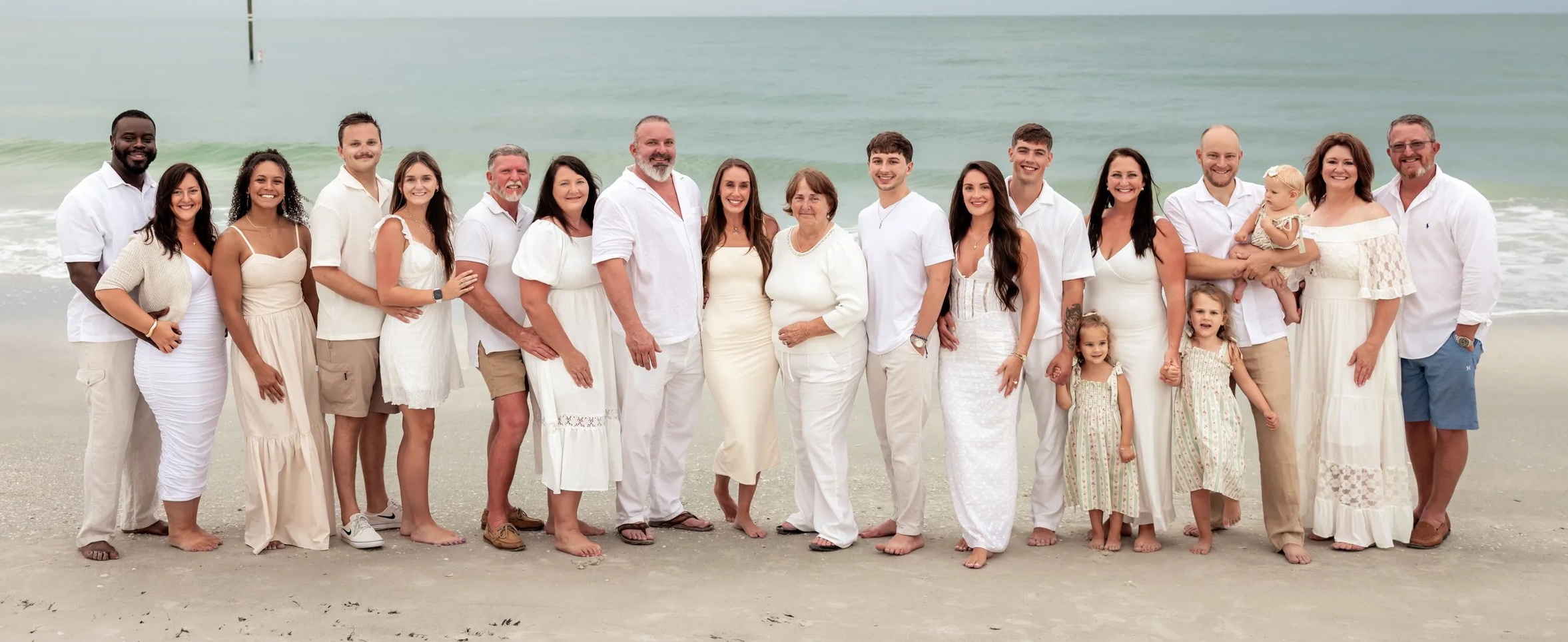 A large multi-generational family dressed in white standing on the beach with the ocean in the background.