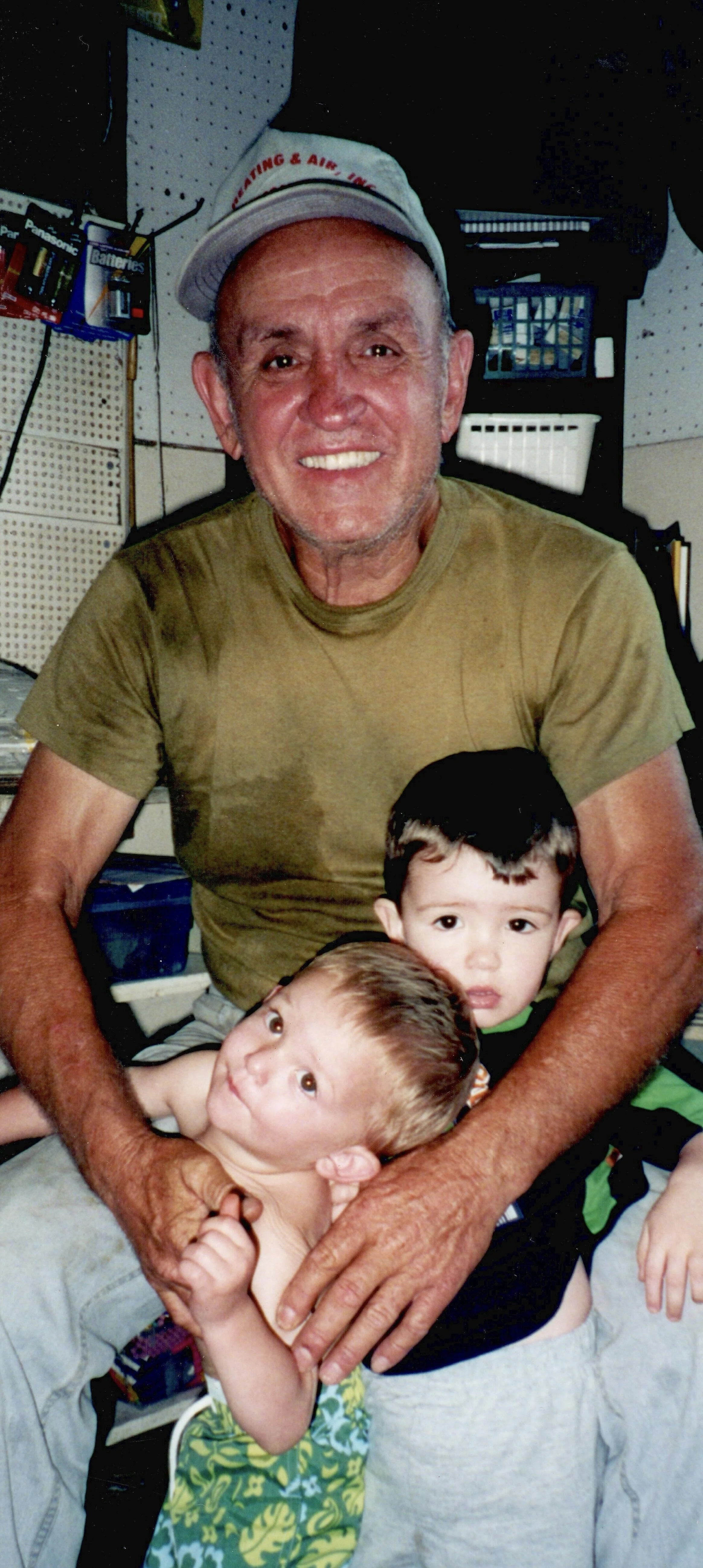 An older man wearing a beige t-shirt and a cap with red lettering is smiling and holding two young boys. The setting appears to be a workshop or hardware store, with shelves displaying batteries and tools in the background.