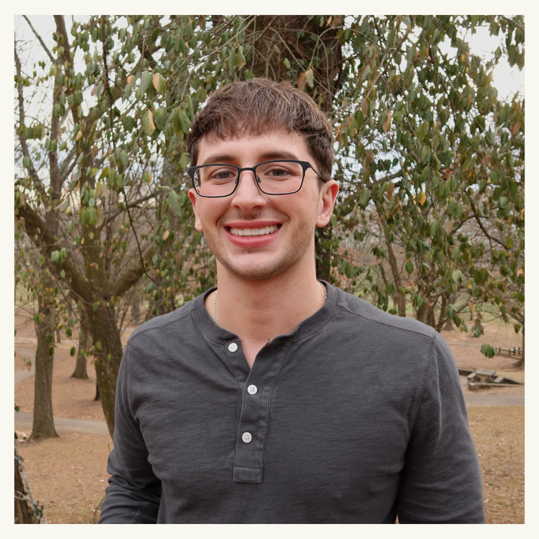 Young man with glasses smiling outdoors, wearing a dark gray shirt, standing in front of a tree with green leaves.
