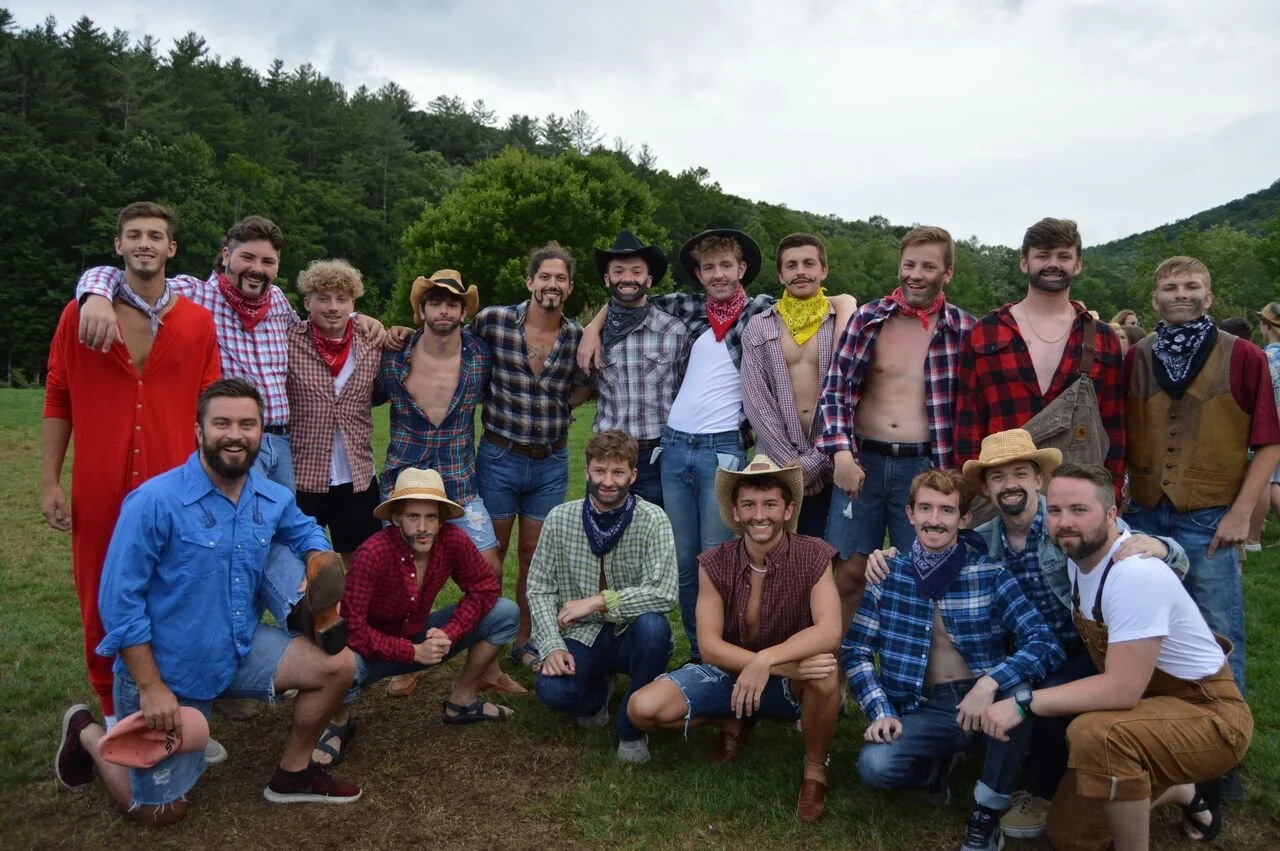 Group of young men dressed in cowboy and western attire, standing outdoors on a grassy field with trees and hills in the background.