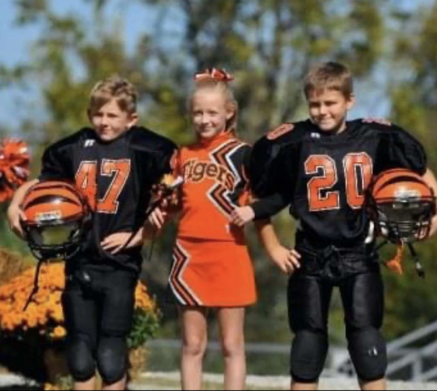 Three young children standing outdoors, two boys in football uniforms holding helmets, and a girl in cheerleading attire standing between them smiling.