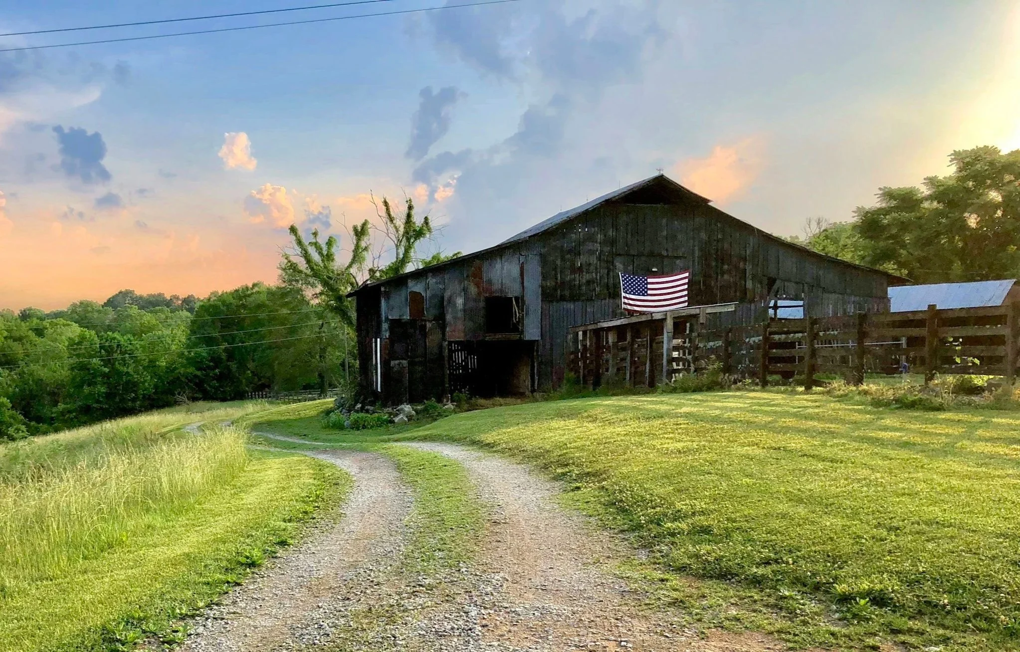 A rustic barn with weathered wood siding and an American flag hanging on the front, situated beside a grassy field with a gravel pathway leading up to it, and surrounded by trees under a partly cloudy sky at sunset.