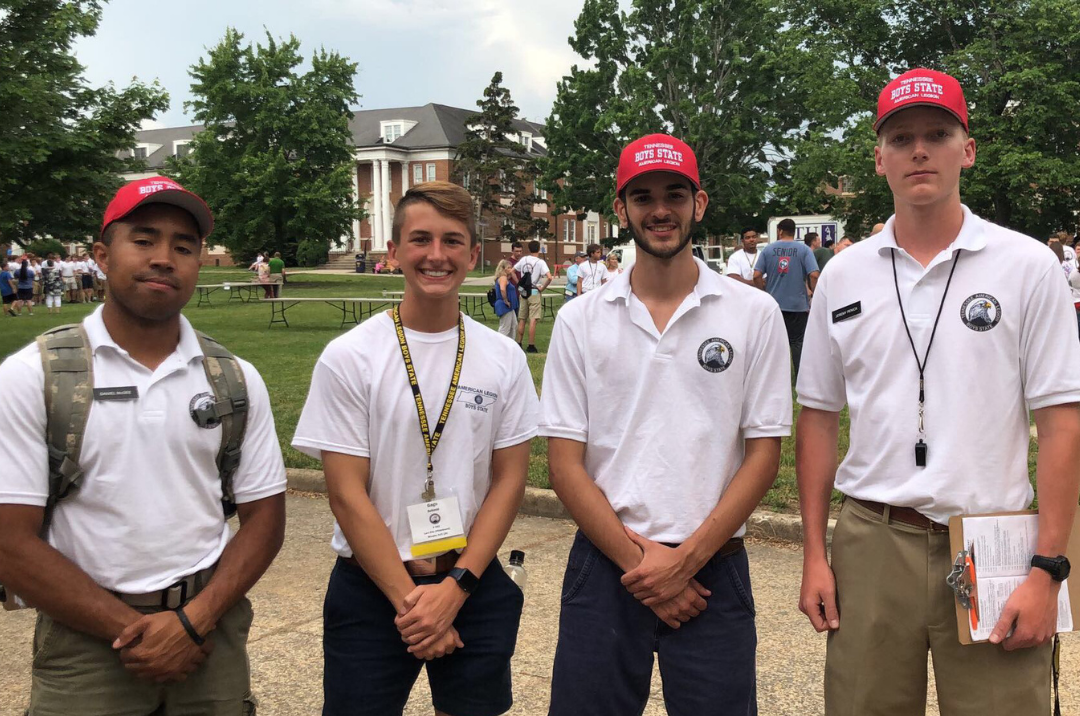 Four young men standing outdoors on a university campus, wearing white polo shirts with logos and red caps that say 'BOYS STATE'. They appear to be participating in a youth leadership event. There are trees, a large building, and other people in the background.
