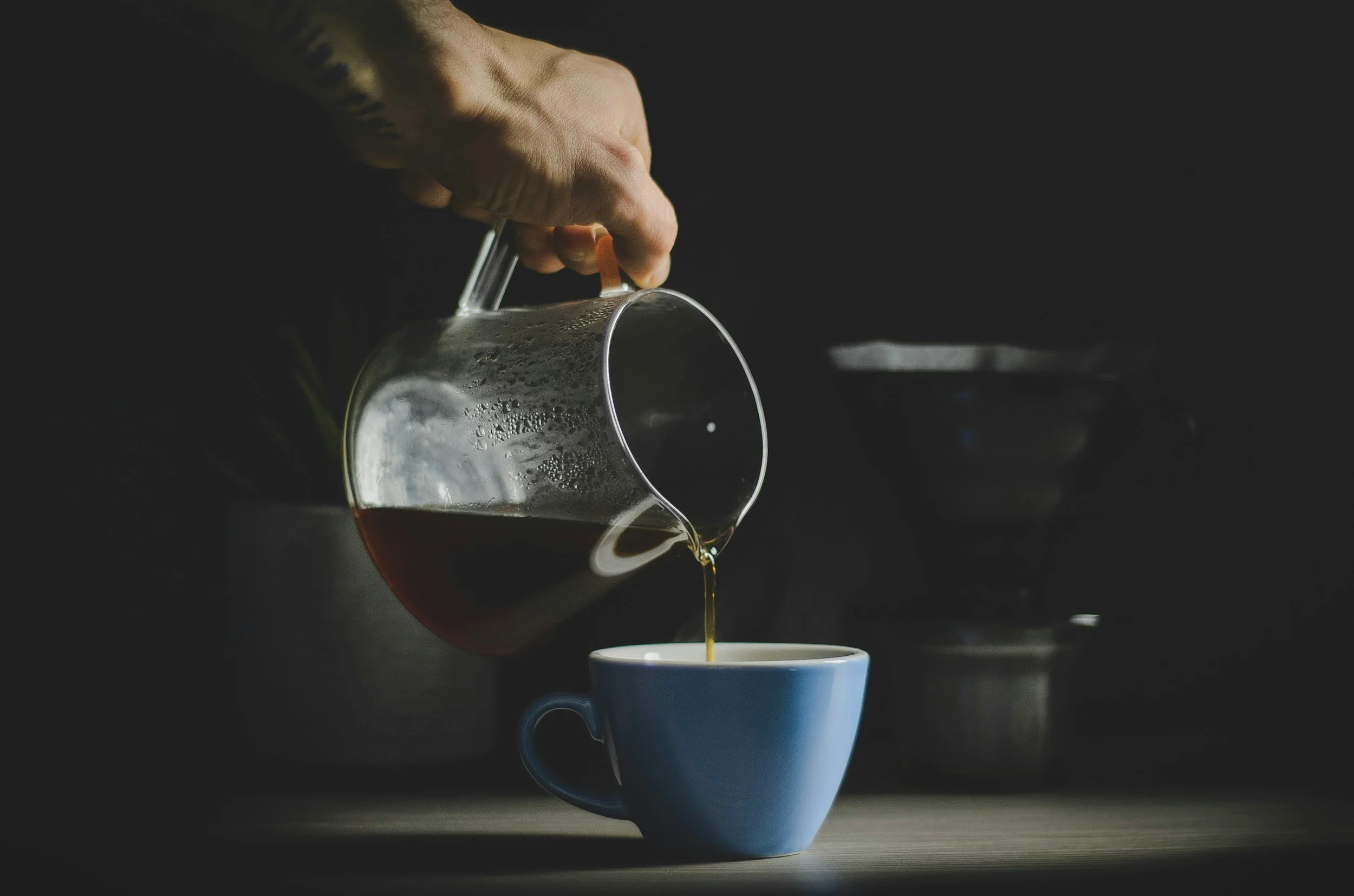 Hand pouring hot coffee from glass pitcher into blue mug.