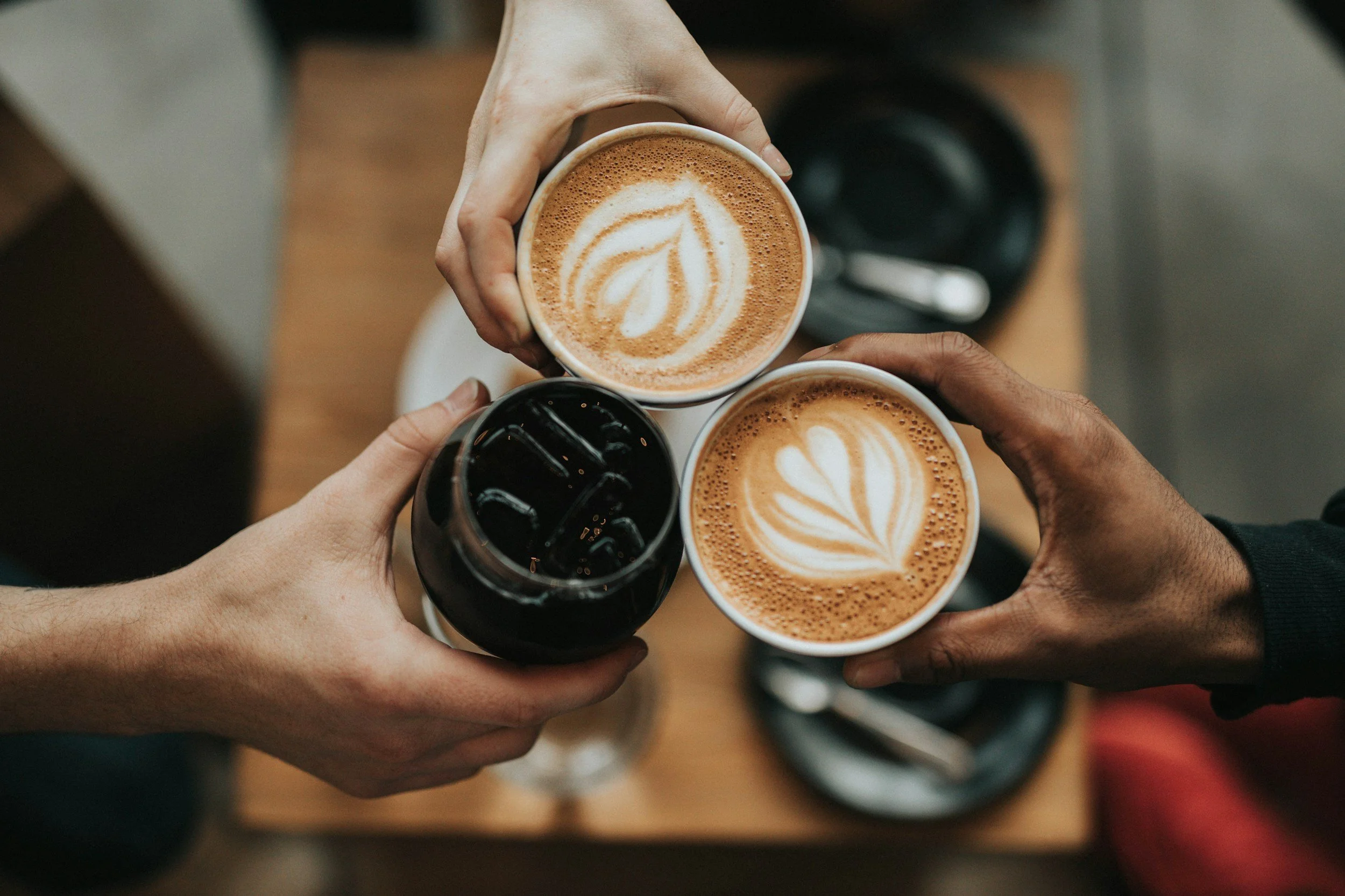 Three different hands holding cups of coffee with latte art and a glass of iced coffee, forming a toast over a wooden table.