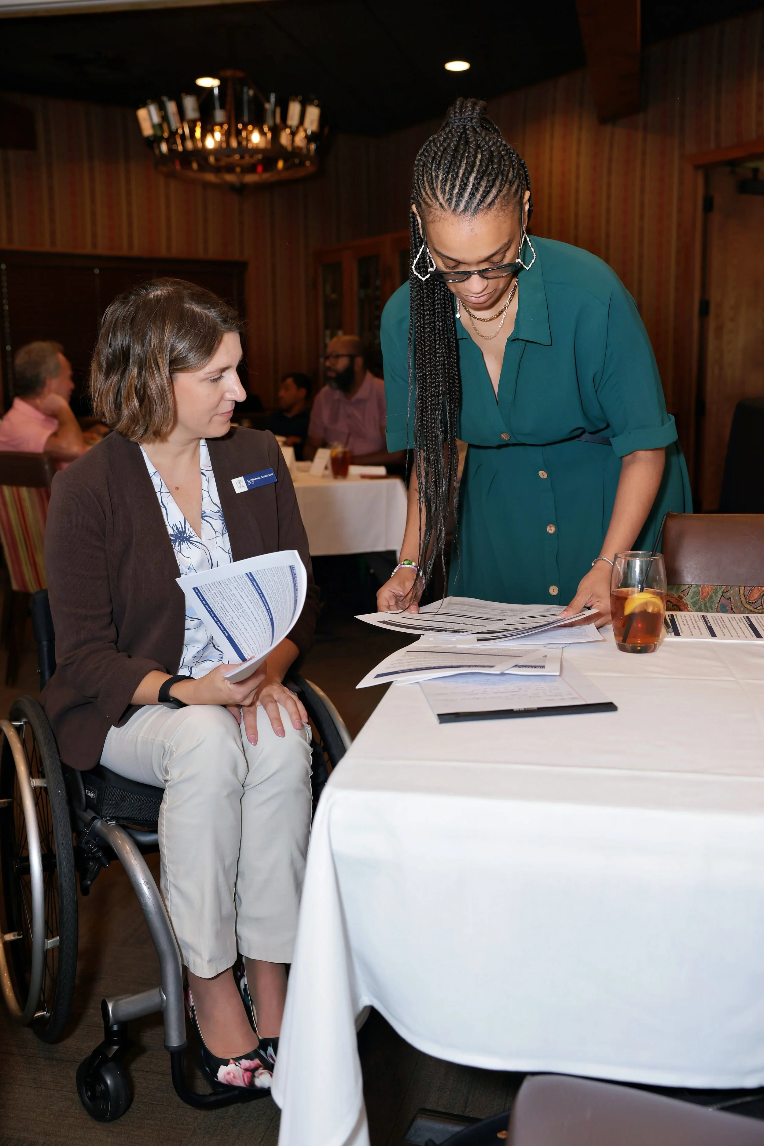 Two women are leading a business luncheon, looking at handouts together. Stephanie - Founder of the Empower Network - is seated in a wheelchair and has short brown hair, Taylor - Board member - is standing next to her.