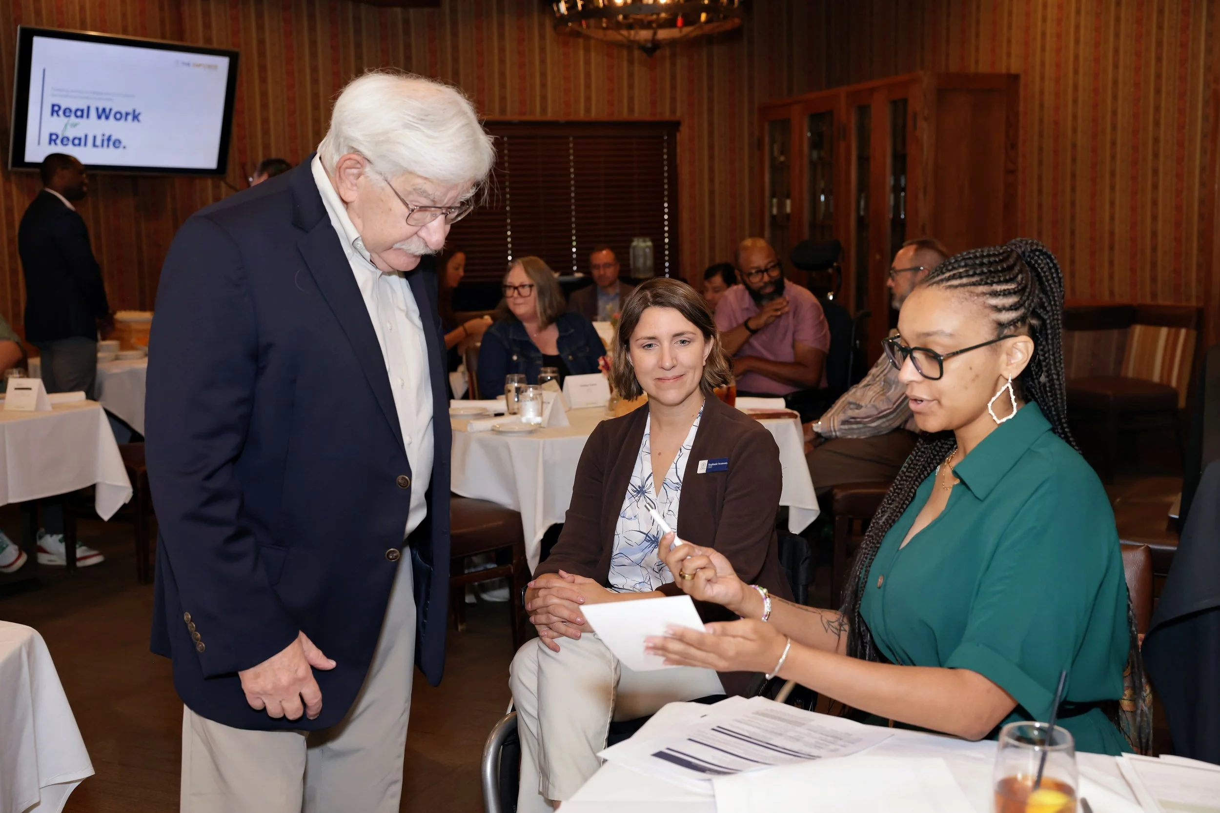 A Black woman in a green dress, a white woman using a wheelchair, and an elderly man with white hair look at a paper together, in a conference room with lunch.