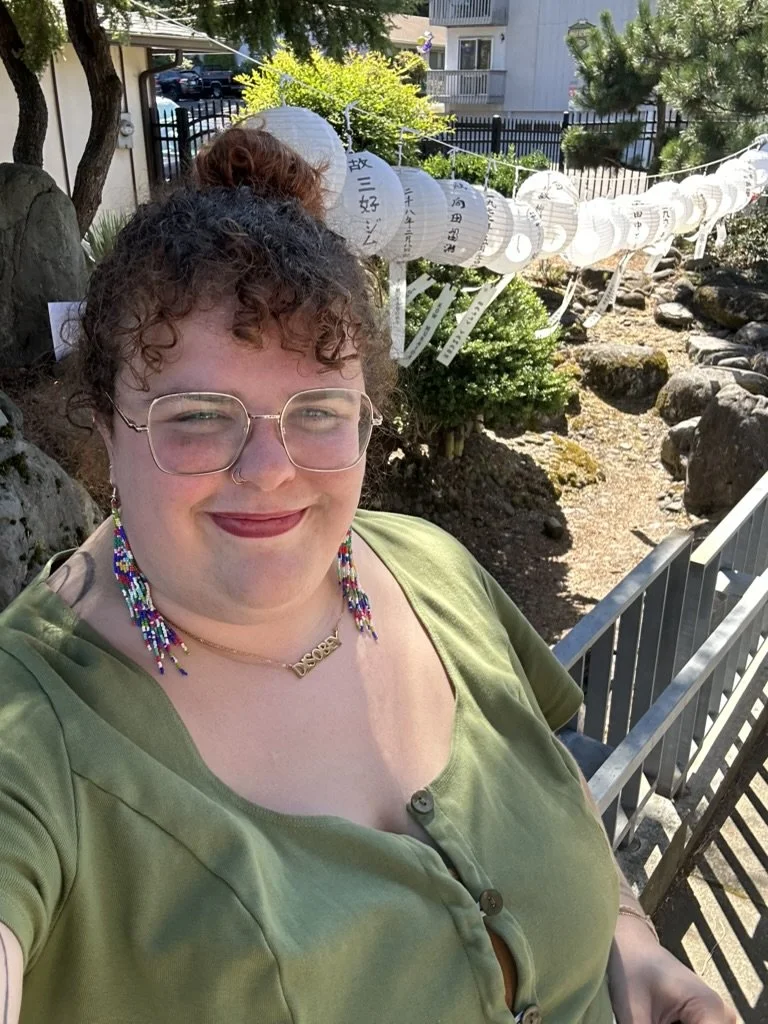 Callie stands outside wearing glasses, earrings, and a green shirt. They are smiling.