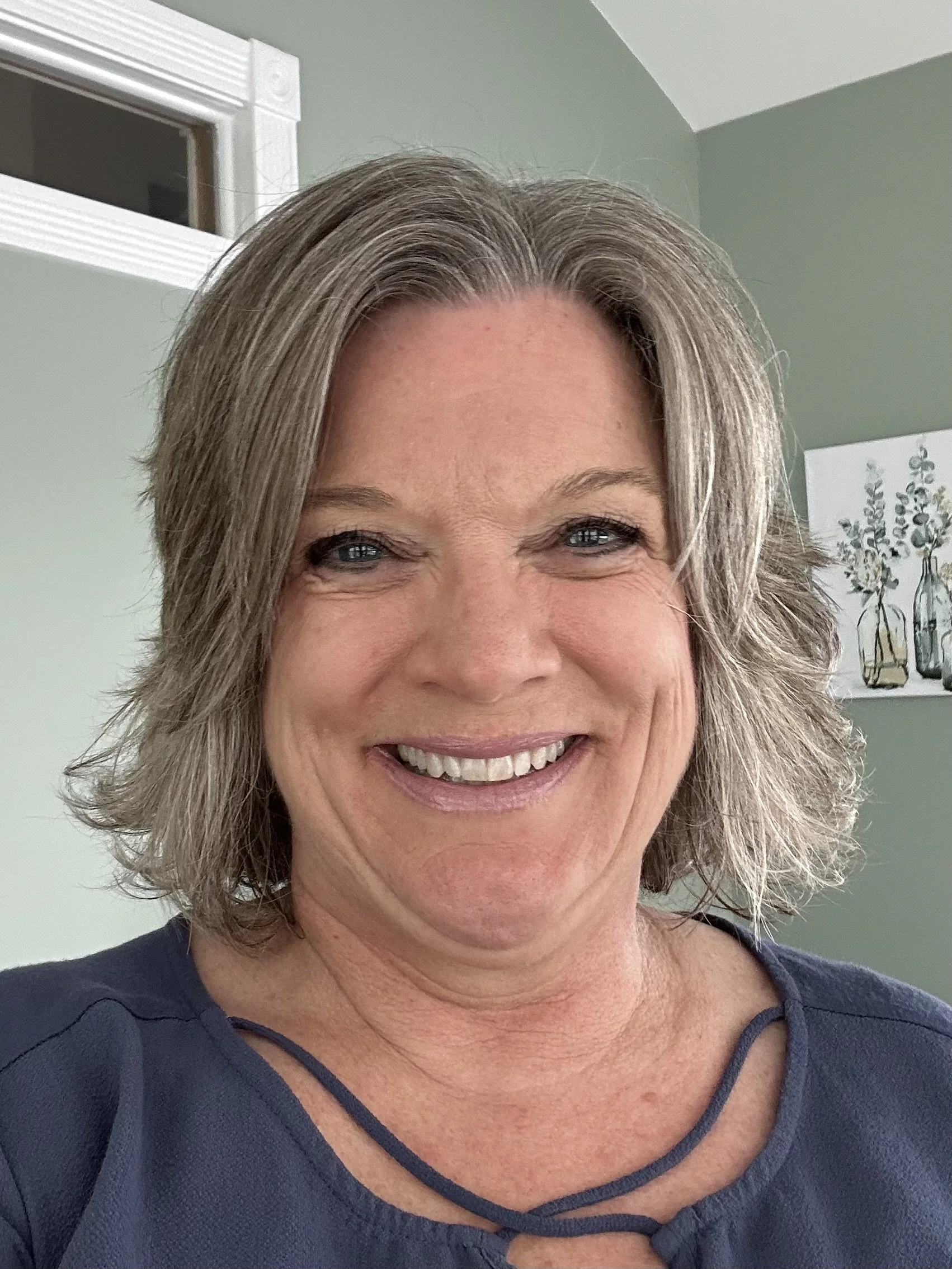 Sue smiles at the camera. She has shoulder-length light or gray hair. She is wearing a slate-blue blouse with a criss cross neckline. Behind them are sage green walls, a small high window, and framed botanical artwork.