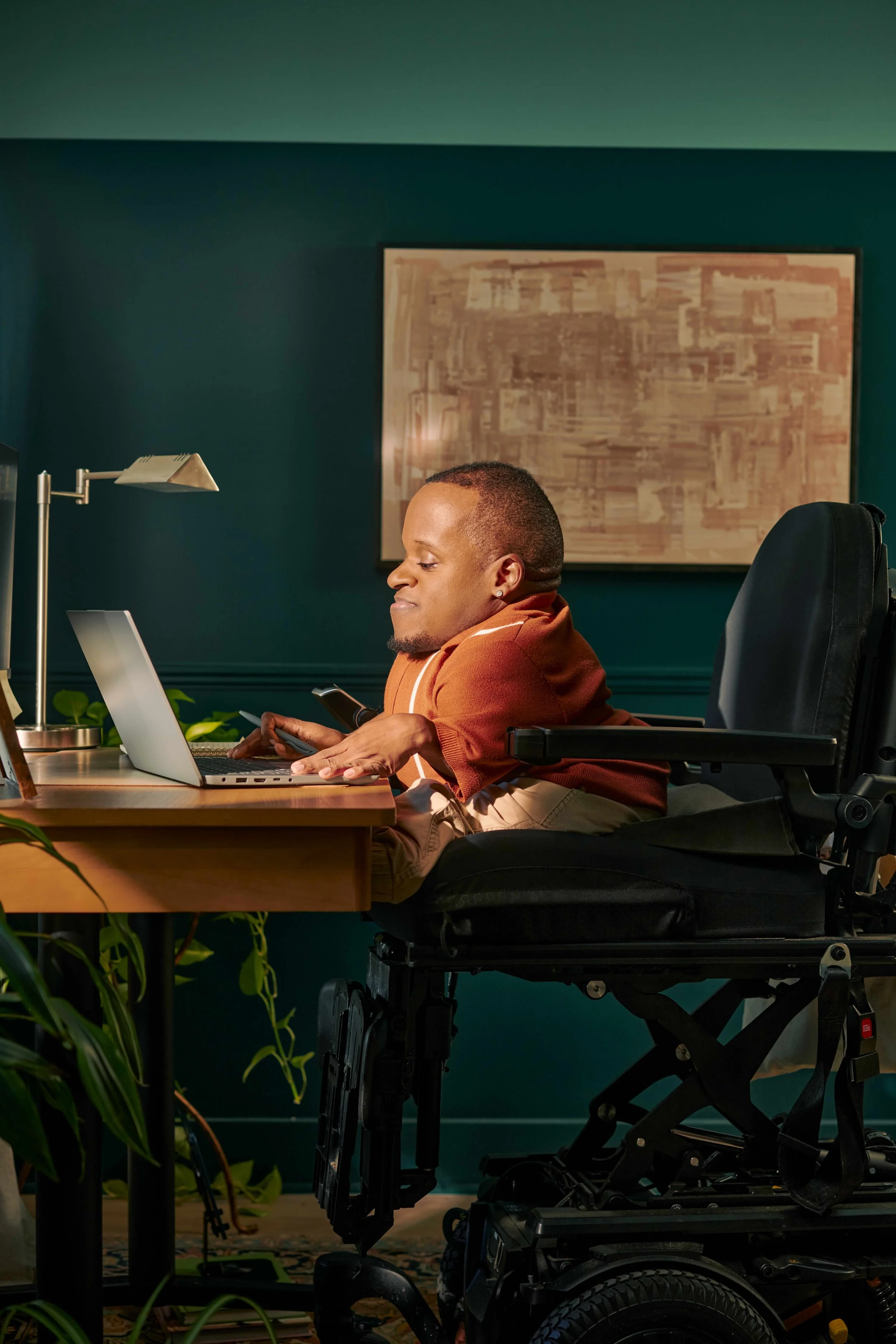 A black man who is a little person works in his home office, seated in his elevated power wheelchair.