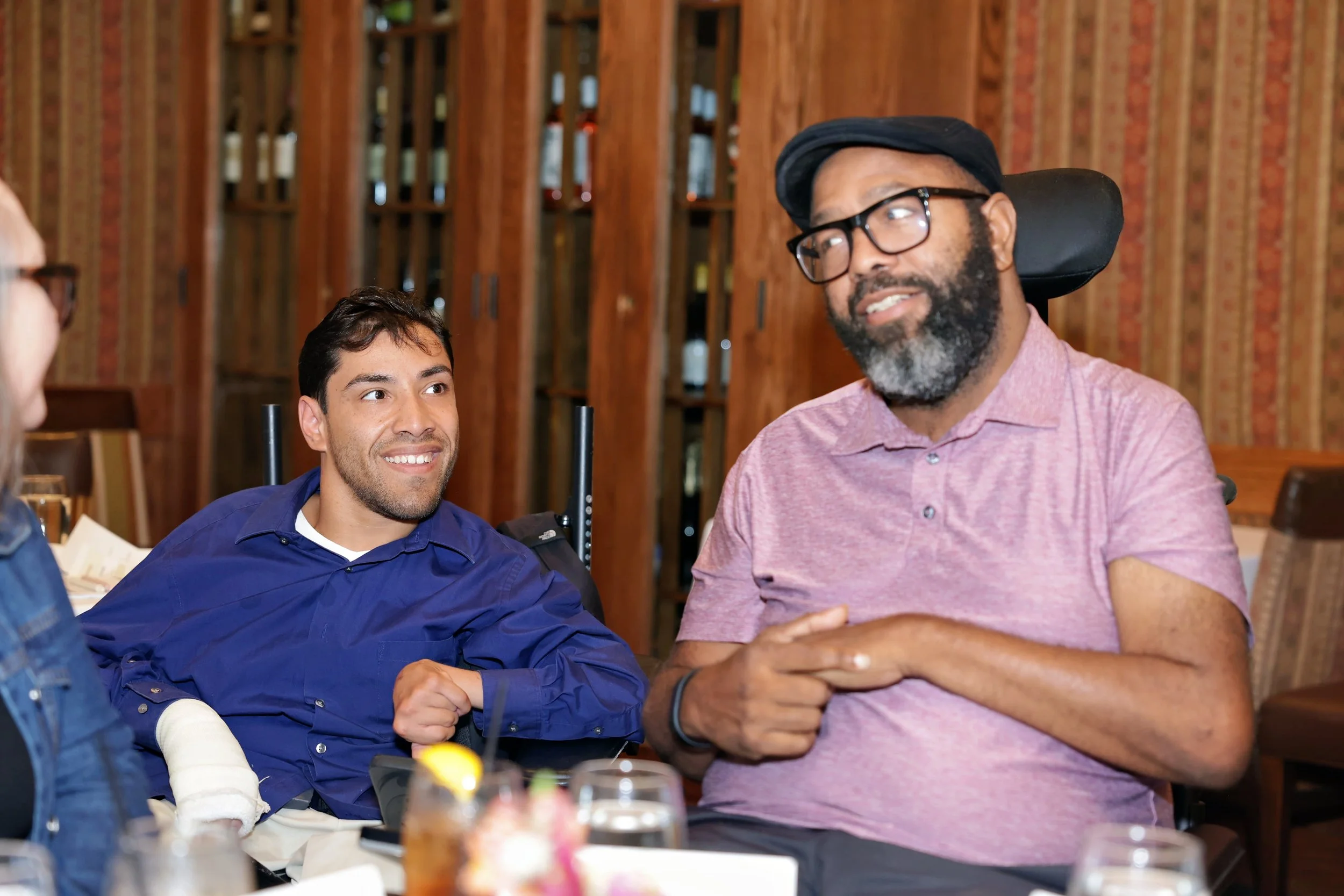 Two men - Ricardo and Brandon - sit in power wheelchairs, smiling and talking at a table.