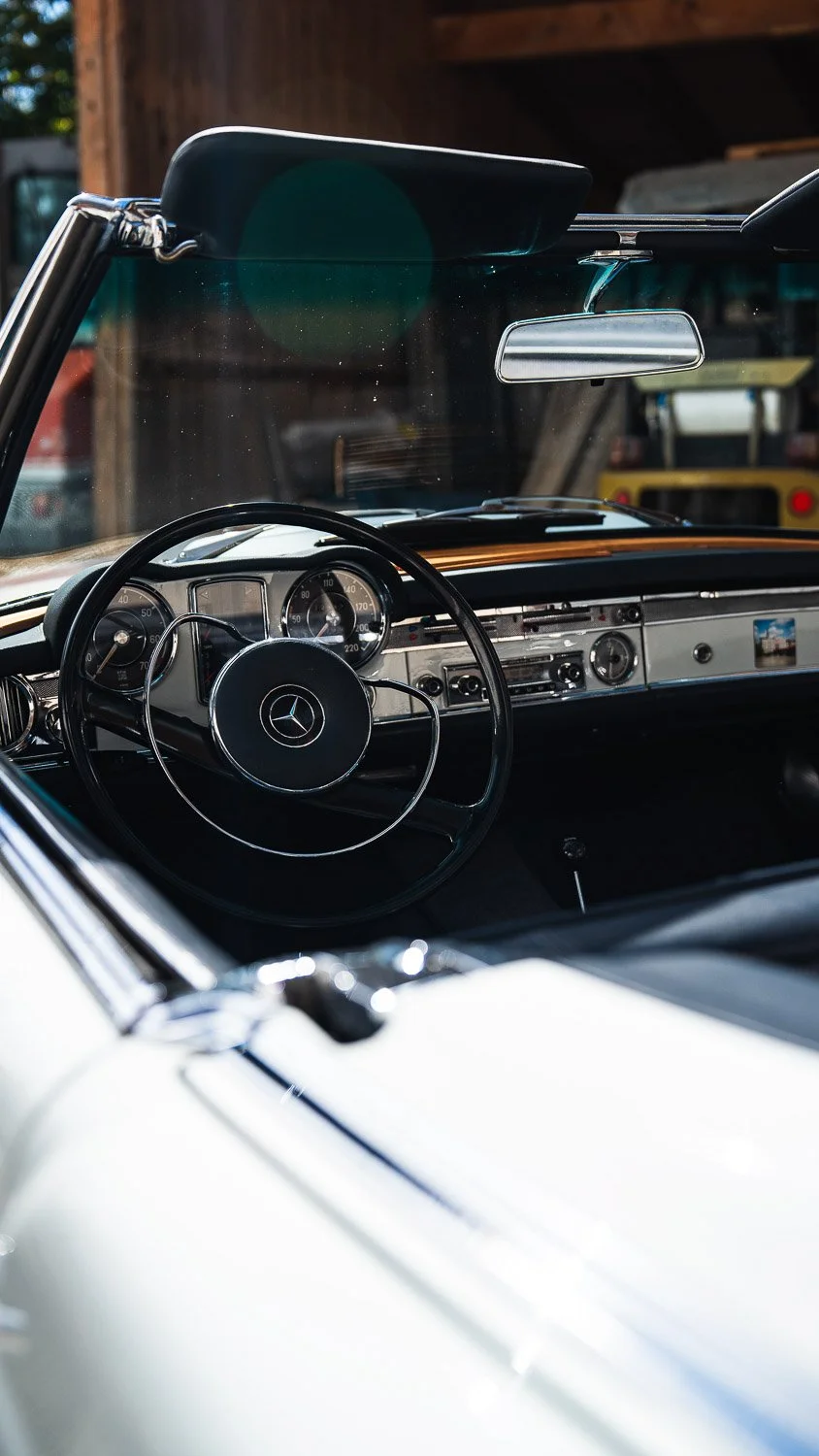 Innenansicht eines klassischen Mercedes-Benz Autos mit schwarzem Lenkrad, Armaturenbrett und Spiegel, geparkt in einer Scheune oder Garage.