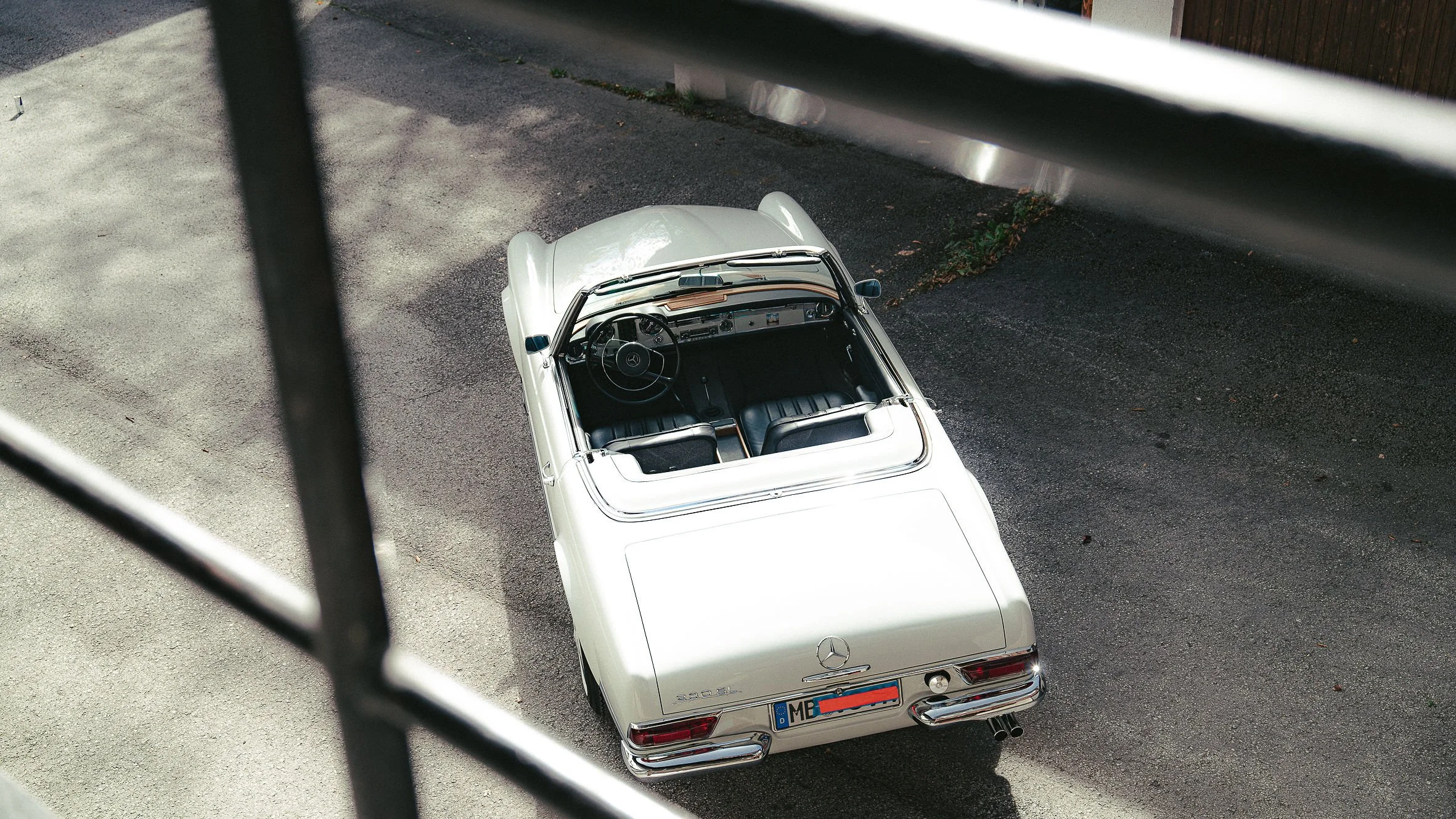 Oberansicht eines weißen klassischen Mercedes-Benz Cabriolets, aufgenommen durch ein Fenster mit Gitter. Das Auto steht auf einer asphaltierte Fläche, die Umgebung enthält einen Gehweg und einen Zaun.