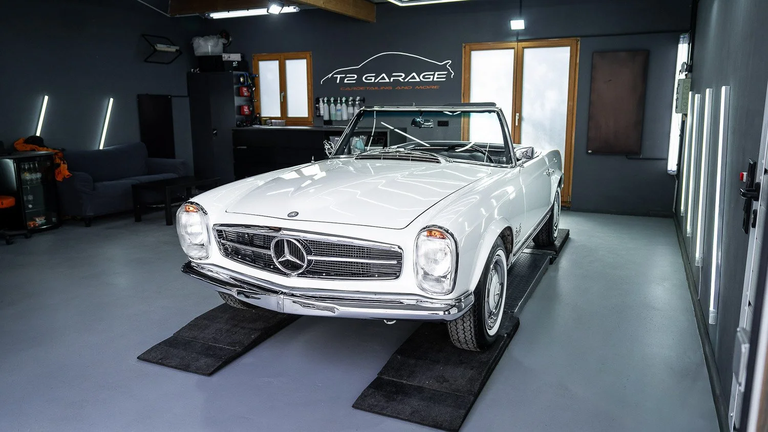 Ein weißer, alte Mercedes-Benz Cabriolet im Autohaus 'T2 Garage', stand auf einer Hebebühne in einem modernen, dunklen Garagenraum.