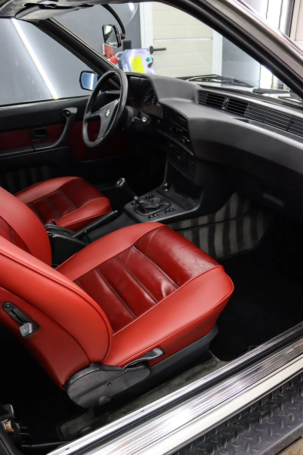 Innenraum eines klassischen Autos mit roter Ledersitzbank, schwarzem Armaturenbrett und Lenkrad, befindet sich im Stellring einer Garage.