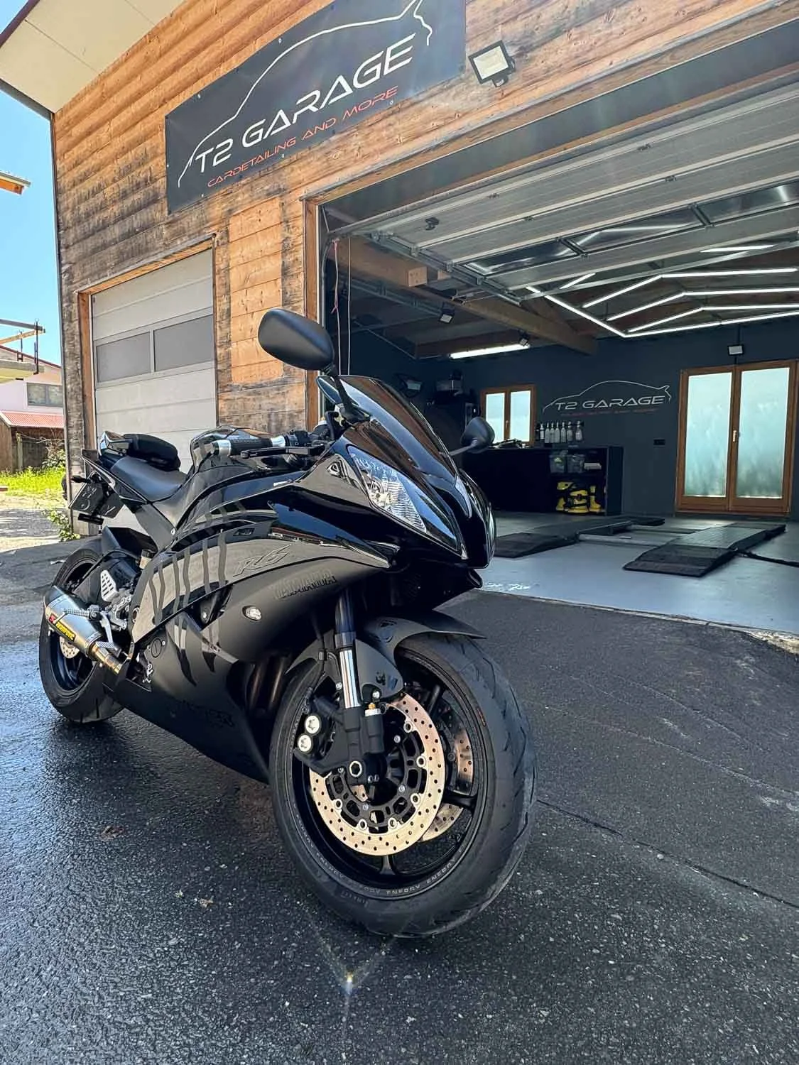 Ein schwarzes Sportmotorrad vor einer offenen Garage mit Holzfassade. Im Hintergrund sind Werkzeuge und Regale zu sehen, sowie ein Logo mit der Aufschrift 'T2 GARAGE' an der Wand.