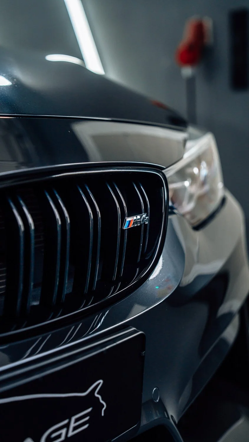 Nahaufnahme des Kühlergrills eines schwarzen BMW M Performance Autos mit M-Logo, im Hintergrund eine weiße Wand und rote Feuerlöscher