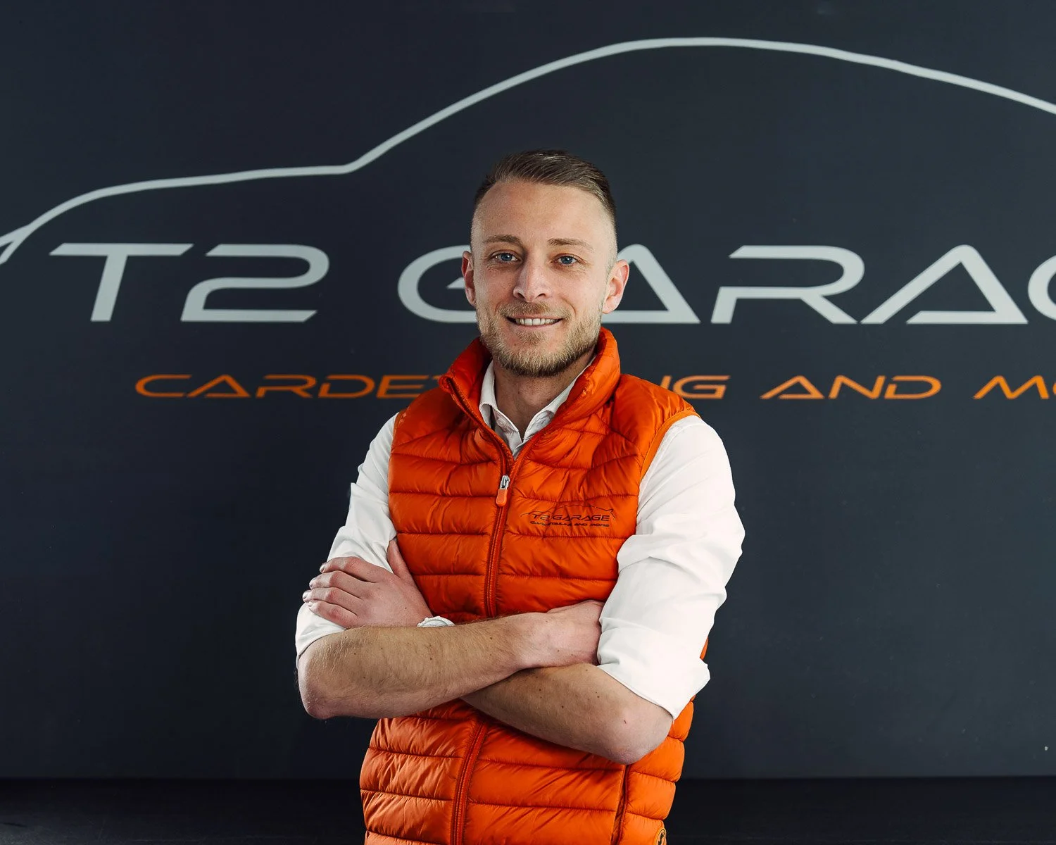 Ein Mann steht vor einem schwarzen Hintergrund mit einem Logo, trägt eine weiße Hemd und eine orange Weste, lächelt, hat die Arme verschränkt.