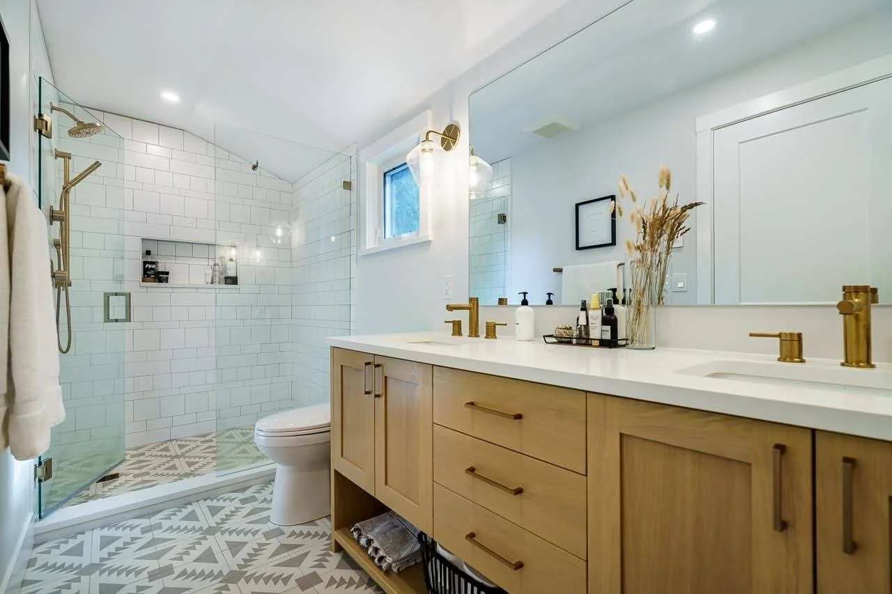 Modern bathroom with a double vanity featuring wooden cabinetry, brass hardware, and a white countertop, along with a large mirror. There is a shower area with glass enclosure, white subway tiles, brass fixtures, and decorative patterned floor tiles inside and outside the shower.