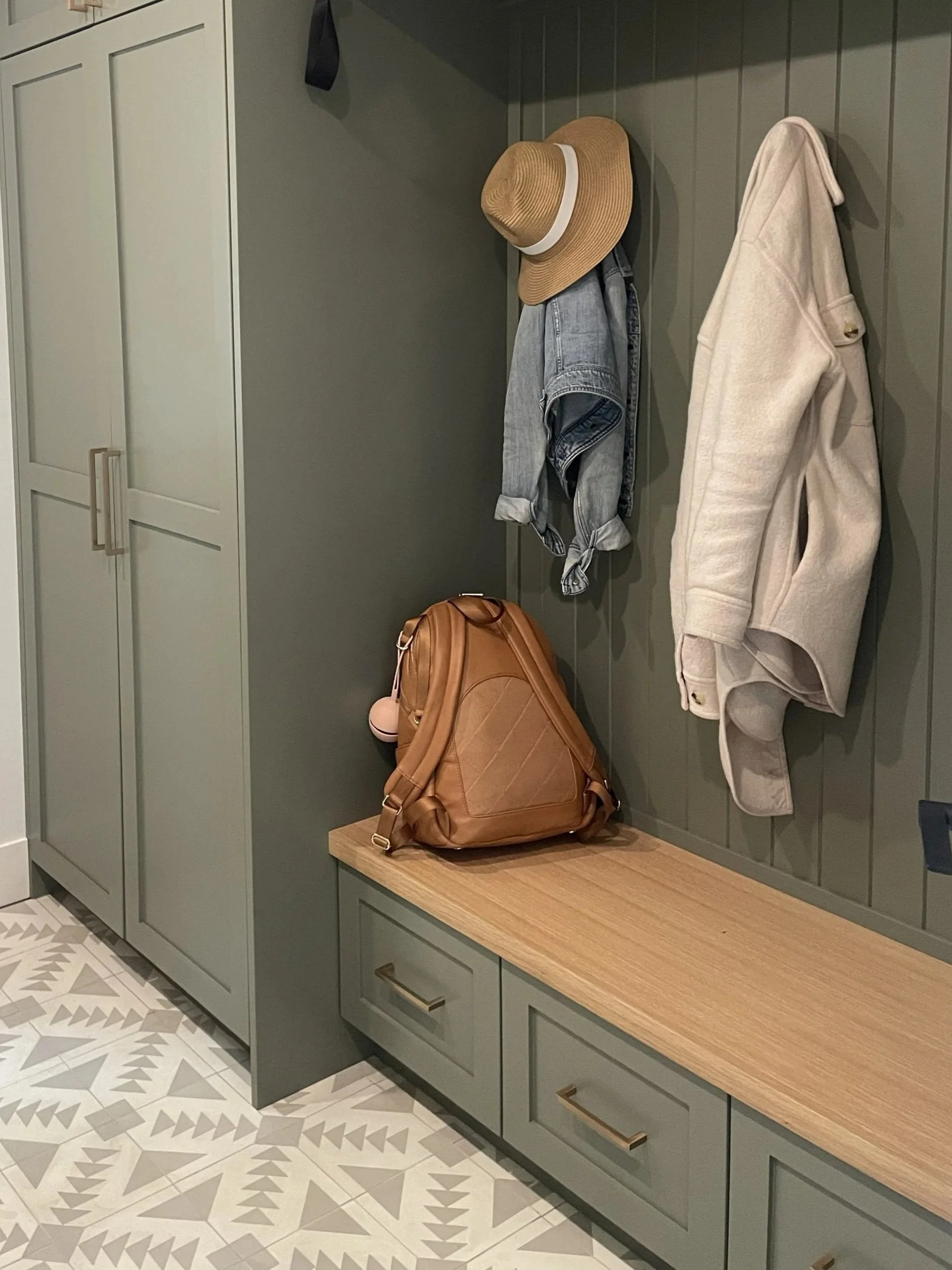 Entryway with a green bench and closet. A straw hat, denim jacket, and beige coat hanging on hooks. A brown backpack on the bench. Patterned tile floor.