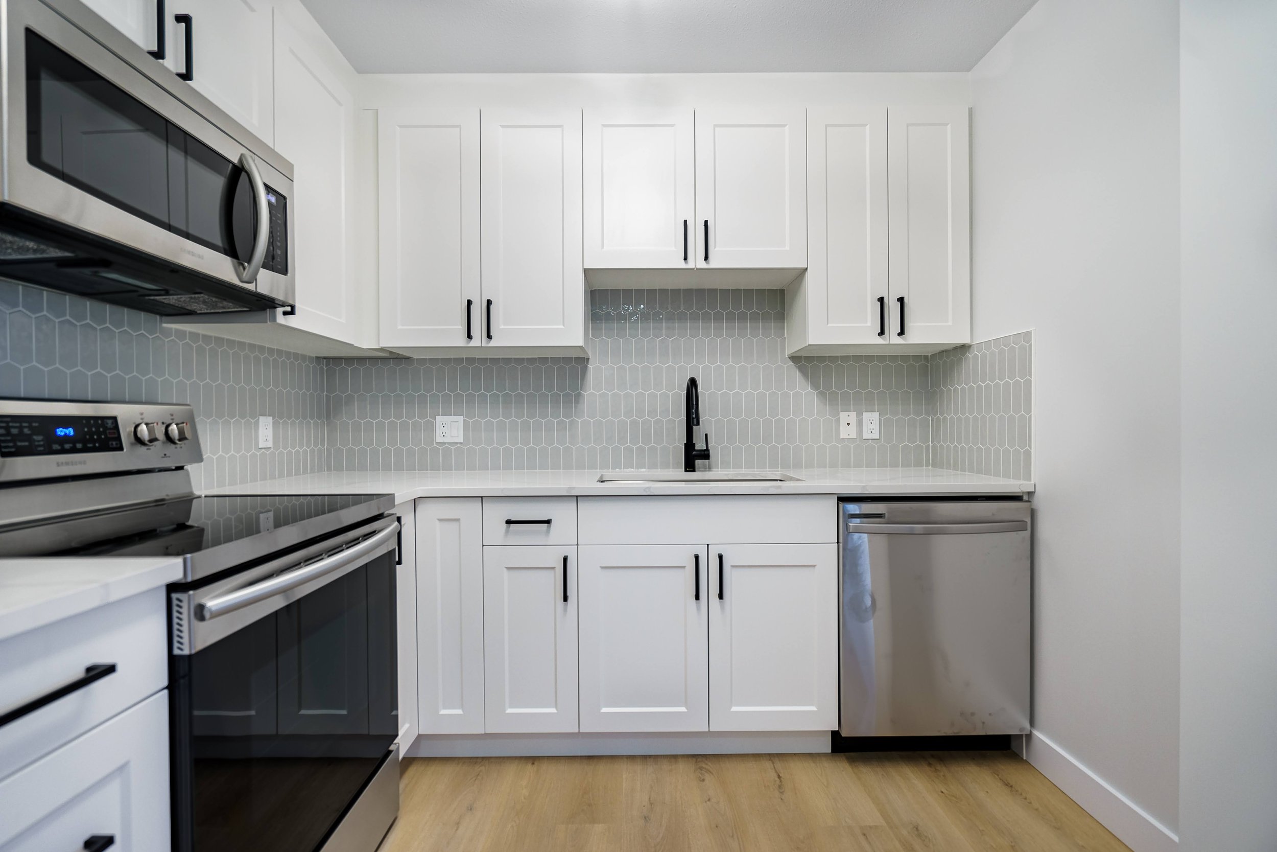 Modern kitchen with white cabinets, gray tiled backsplash, black hardware, stainless steel appliances including microwave, oven, and dishwasher, and a black faucet over the sink.