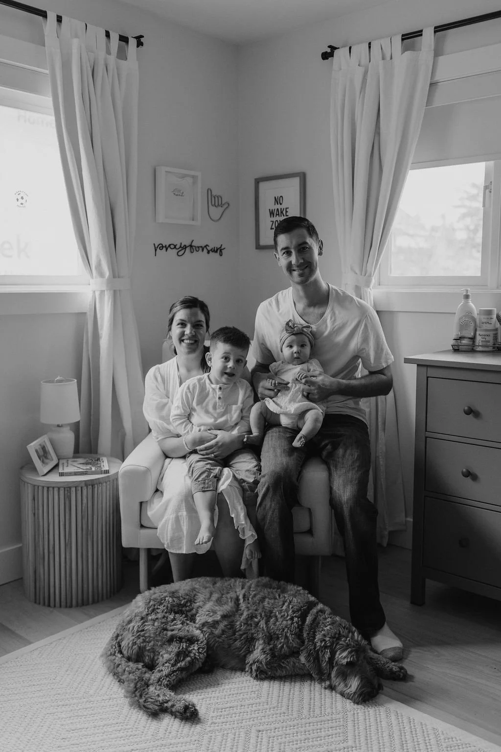 A happy family of four with a dog sitting on a rug, inside a cozy living room with white curtains, framed artwork, and a side table with books and a lamp.