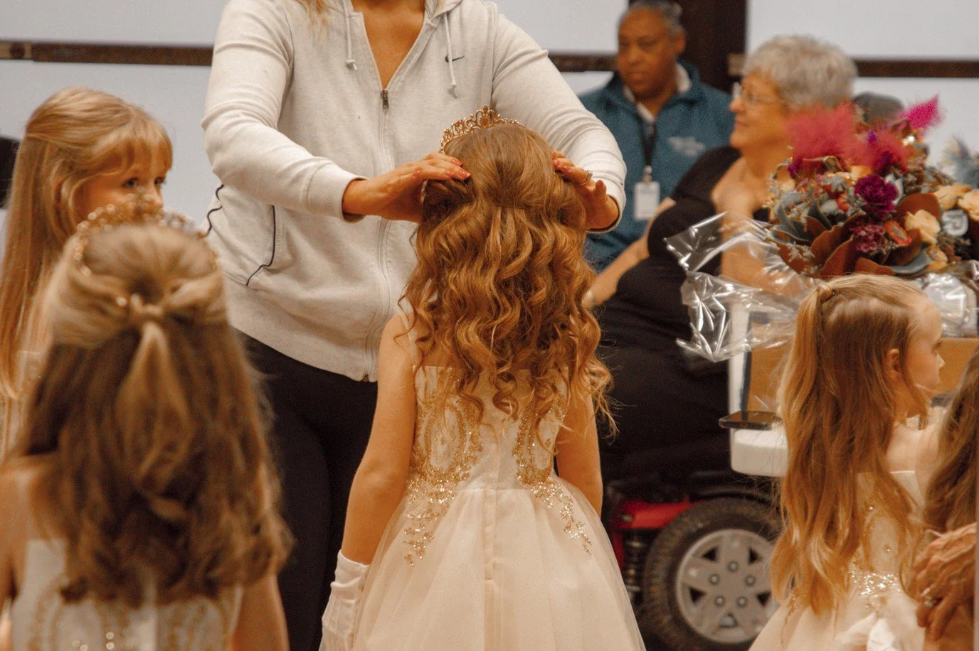 A young girl with long, curly, red hair wearing a white dress is getting a tiara placed on her head by an adult woman. Several other young girls with similar dresses and hair are nearby, and in the background, there are older women and a large bouque