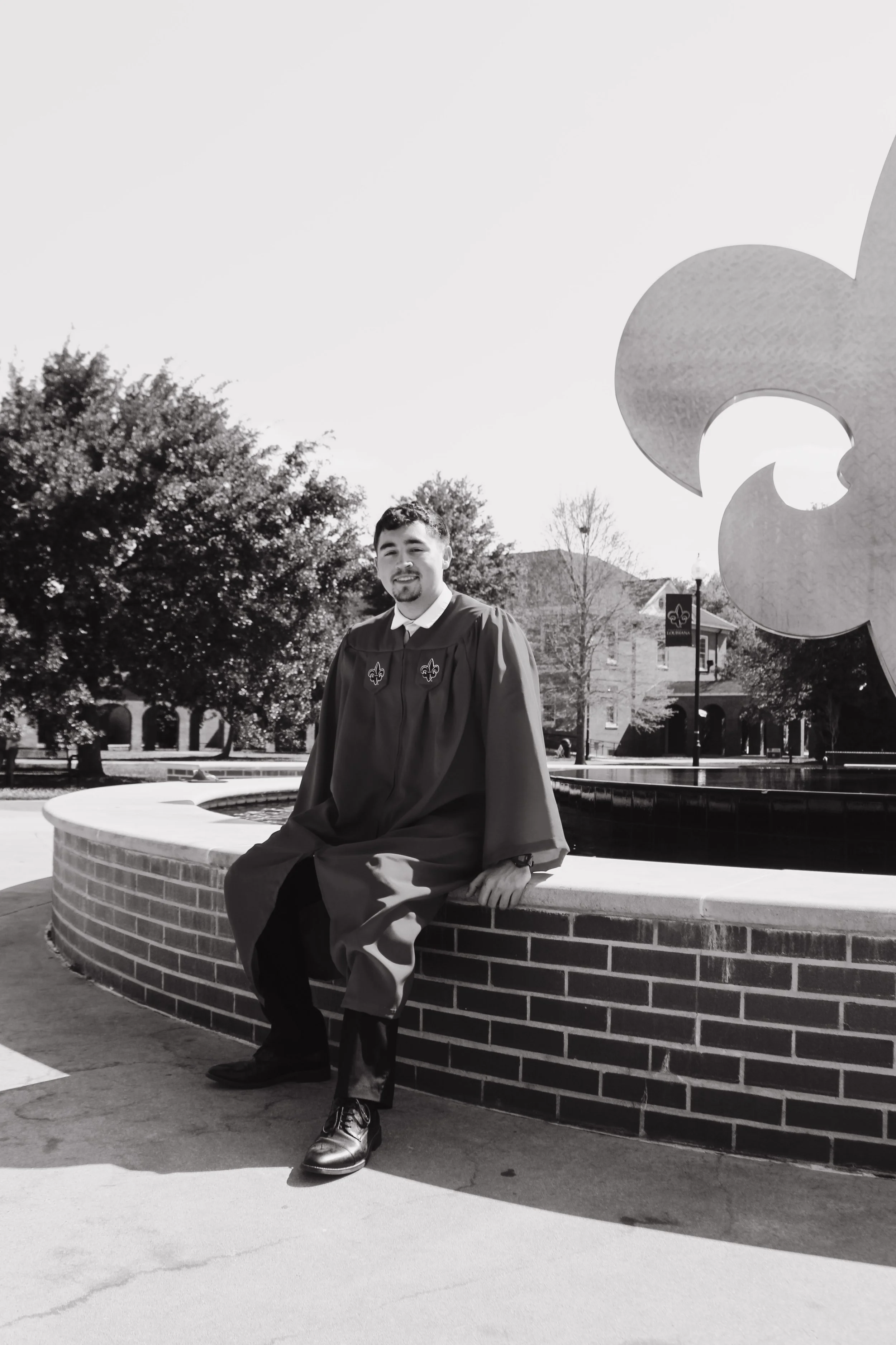 A young man in a graduation gown and cap sitting on a brick circular bench in an outdoor plaza, with trees and a building in the background.