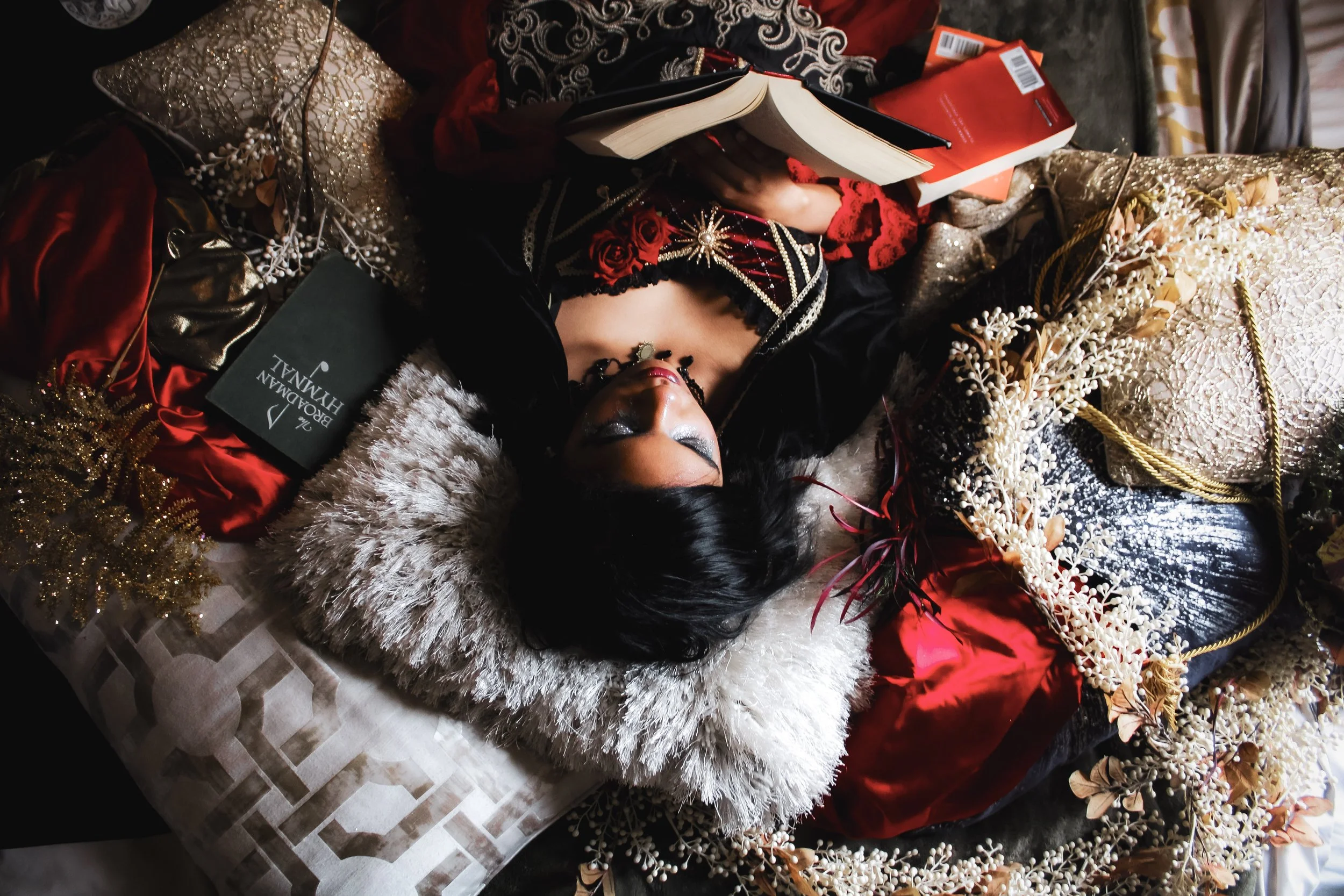 A woman is lying down surrounded by luxurious fabric, gold and white decor, and various books, with her eyes closed and makeup on.
