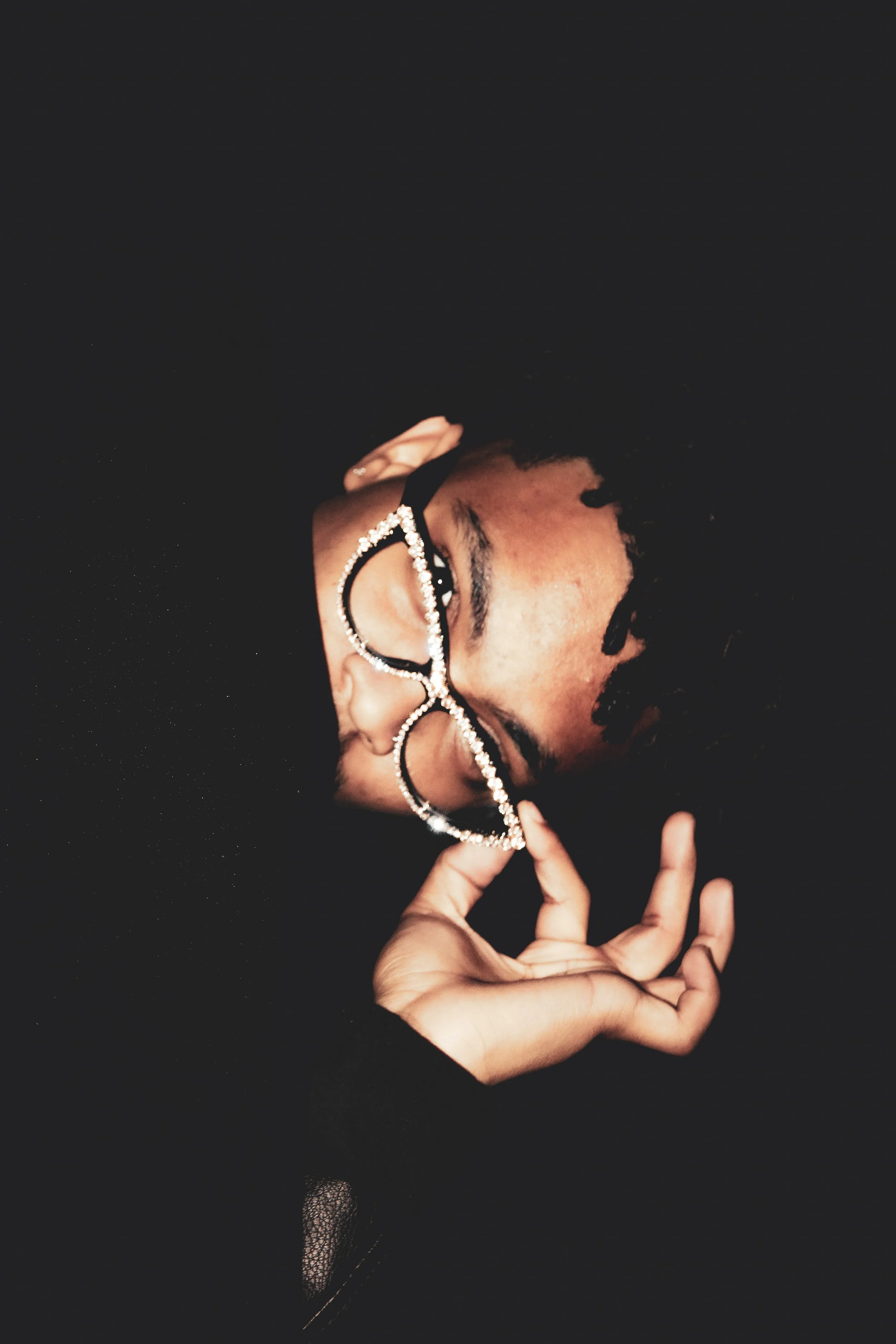 A person with glasses that have rhinestones, holding them with one hand, lying on a dark surface, smiling, with dreadlocks and wearing black clothing.