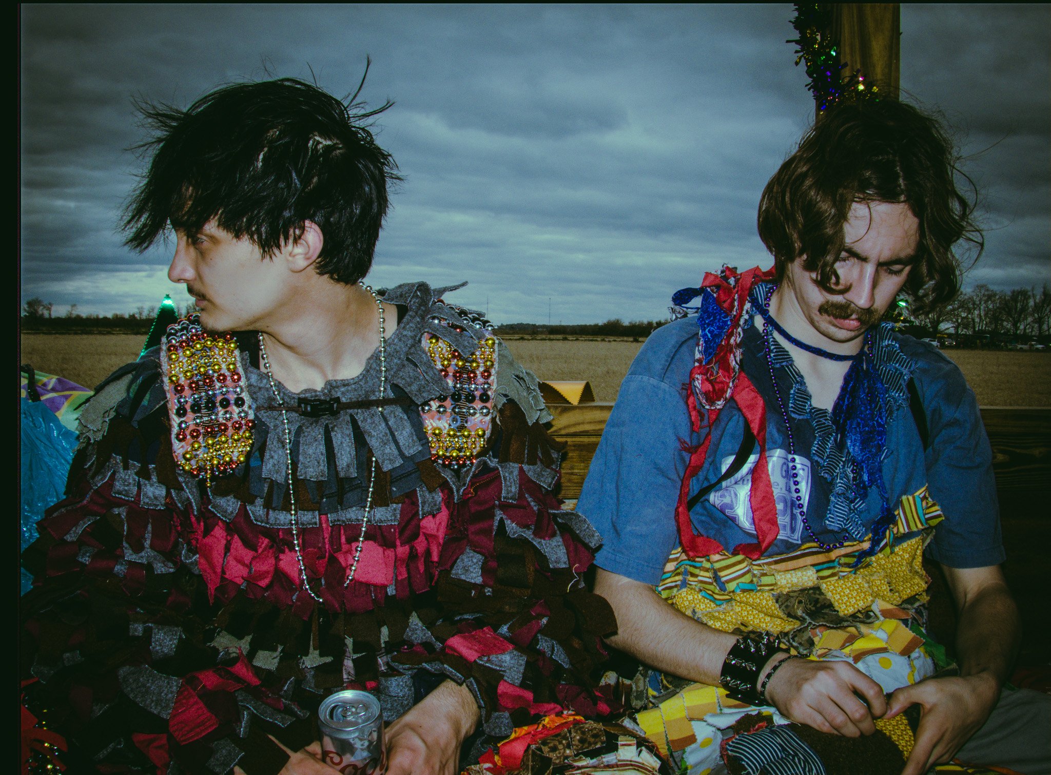 Two men with dark, messy hair and facial hair sitting outdoors on a cloudy day. One wears a costume decorated with beads and fabric patches, and the other wears a blue shirt with a red bandana and patterned fabric. They are engaged with colorful fabric pieces in front of them.
